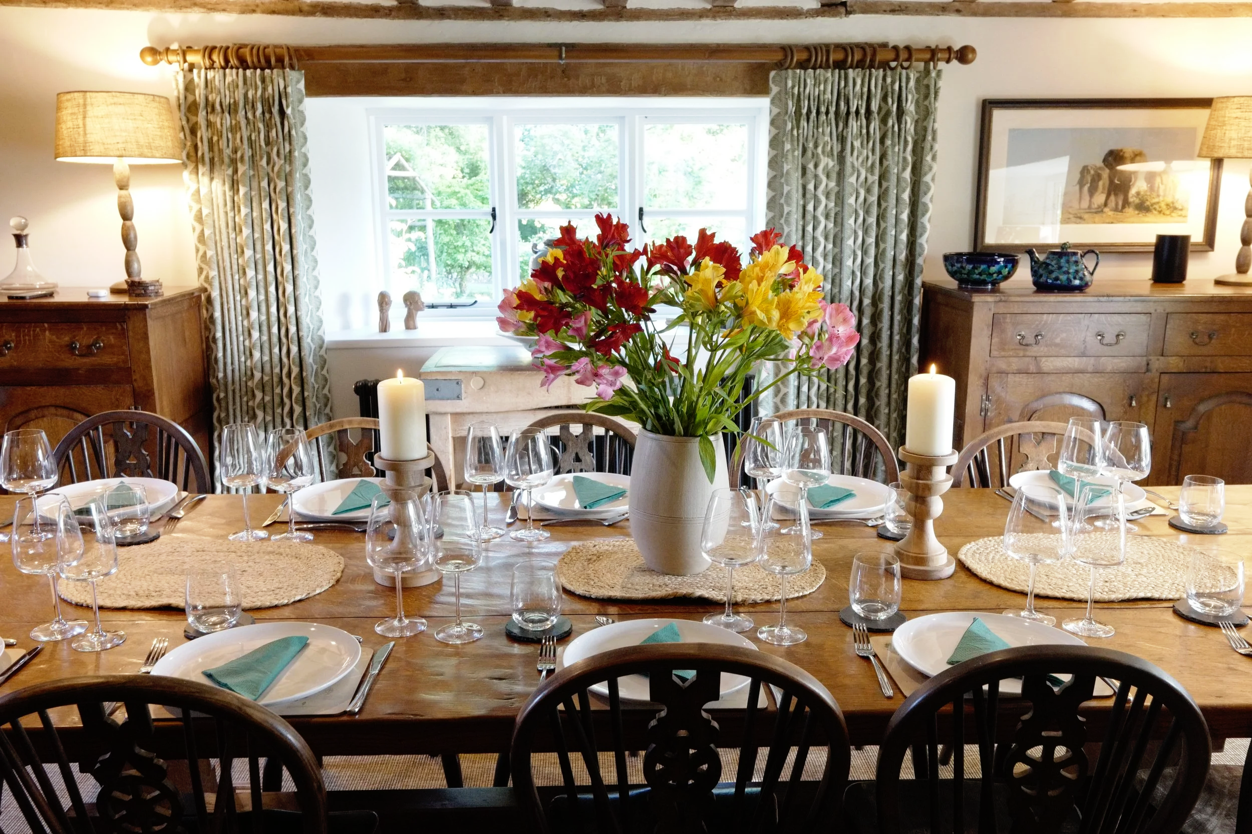 A dining room with a large wooden table set with wine glasses, white plates, and green napkins for a meal. There is a large vase with red, yellow, and pink flowers in the center of the table. The room has a window with patterned curtains, decorative 
