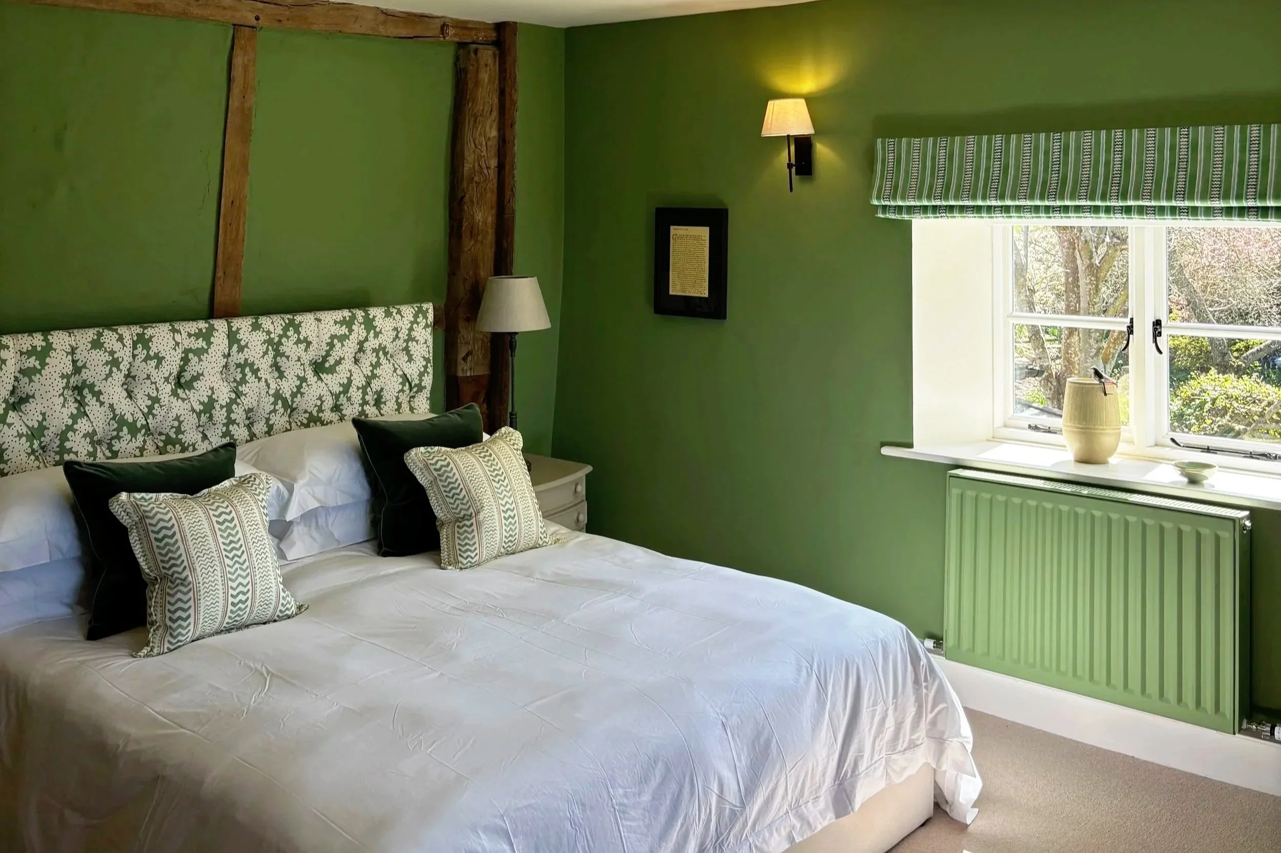 A bedroom with green walls and a white bed featuring multiple pillows with striped and holly patterns. There is a bedside lamp, a framed document, and a window with a green and white striped valance. The window shows trees outside, and a small table 