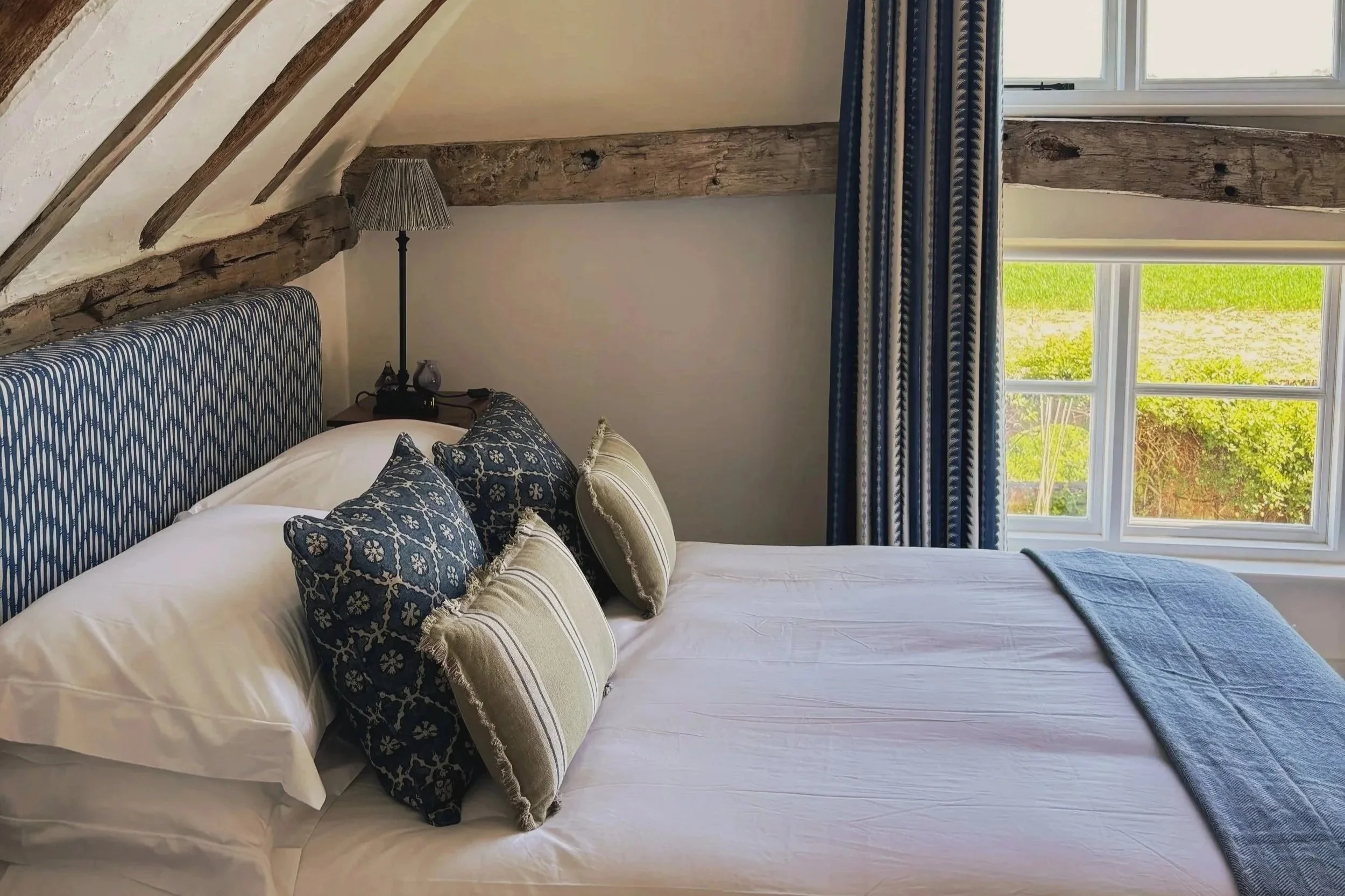 A cozy bedroom with a bed featuring white linens and patterned pillows, wooden beams on the ceiling, a window with blue curtains showing greenery outside, and a small bedside lamp.