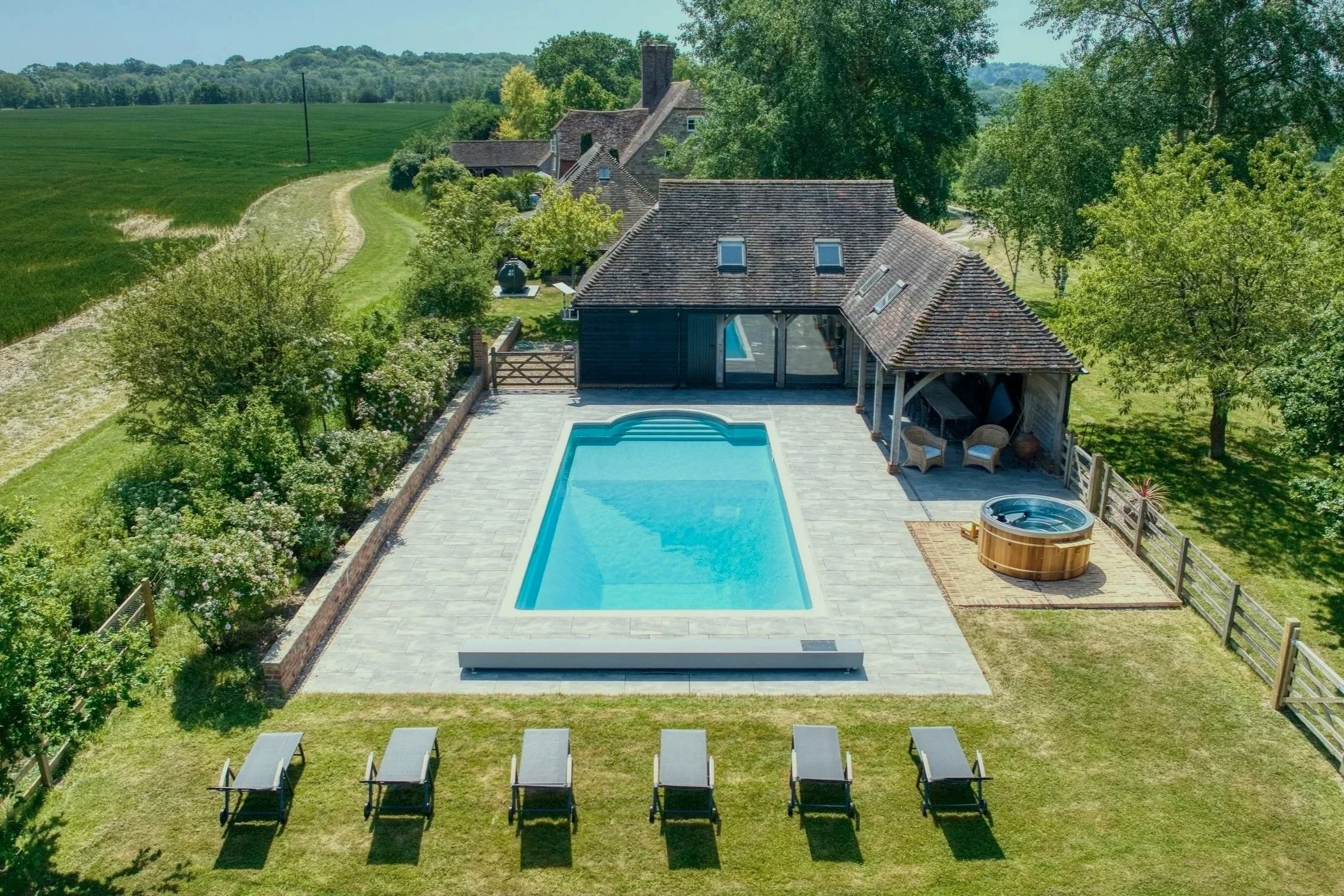 Aerial view of a backyard with a swimming pool, hot tub, and lounge chairs, surrounded by trees and fencing, with a house and lush green fields in the background.