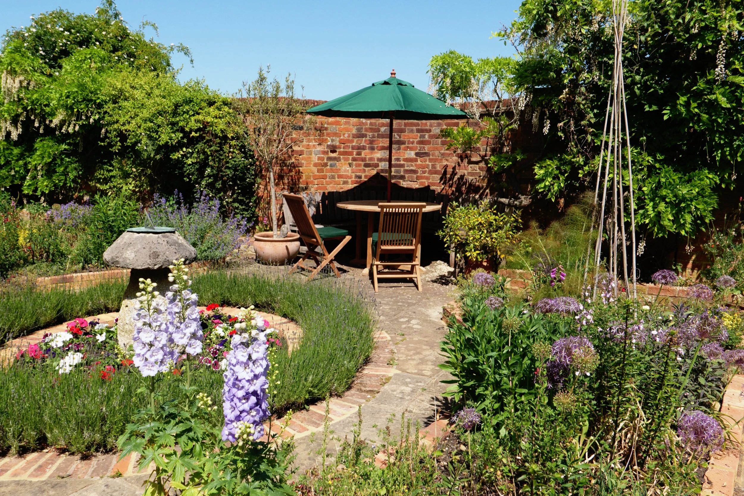 A lush garden with colorful flowers, green shrubs, and trees. A stone pathway leads to an outdoor seating area with a round table, wooden chairs, and a large green umbrella. A brick wall with flowering vines is in the background.