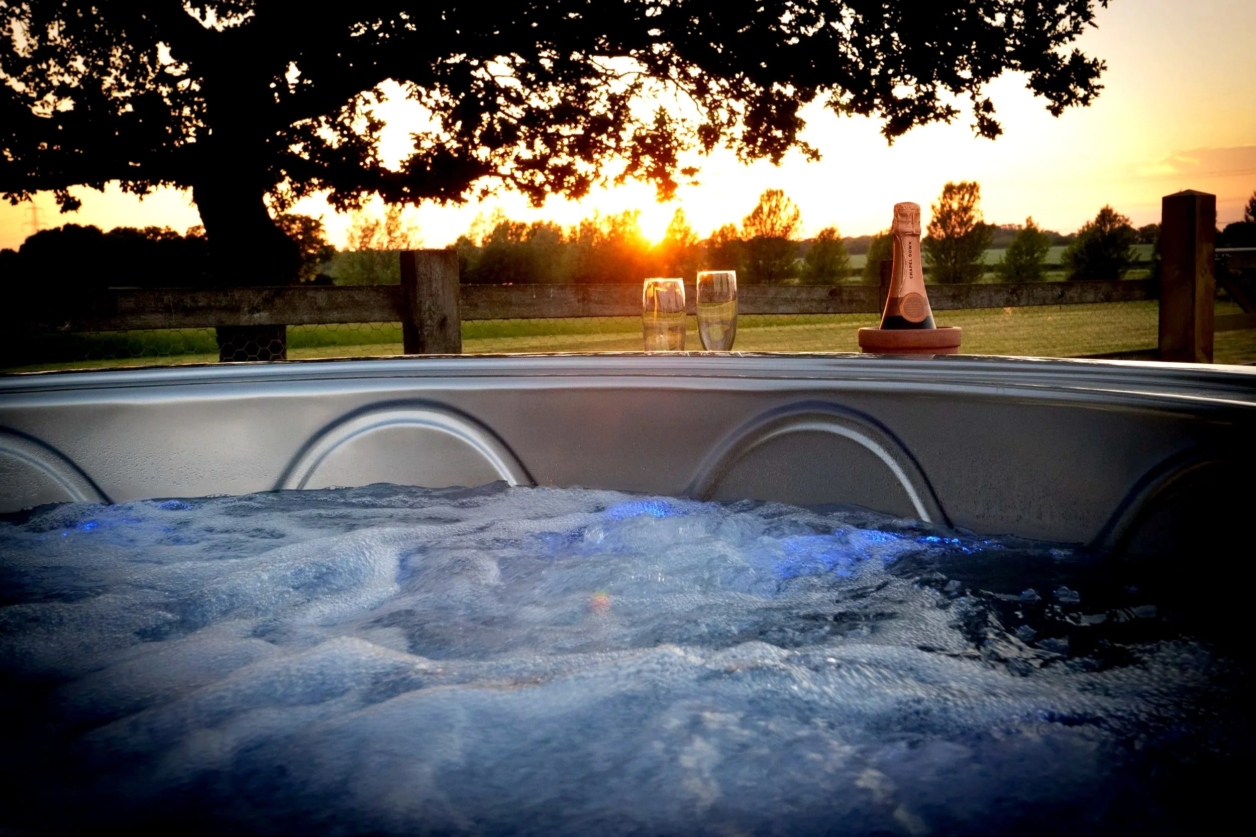 A hot tub with bubbling water, a bottle of sparkling beverage in a terracotta holder, and two glasses on the edge, set outdoors at sunset with trees and a wooden fence in the background.