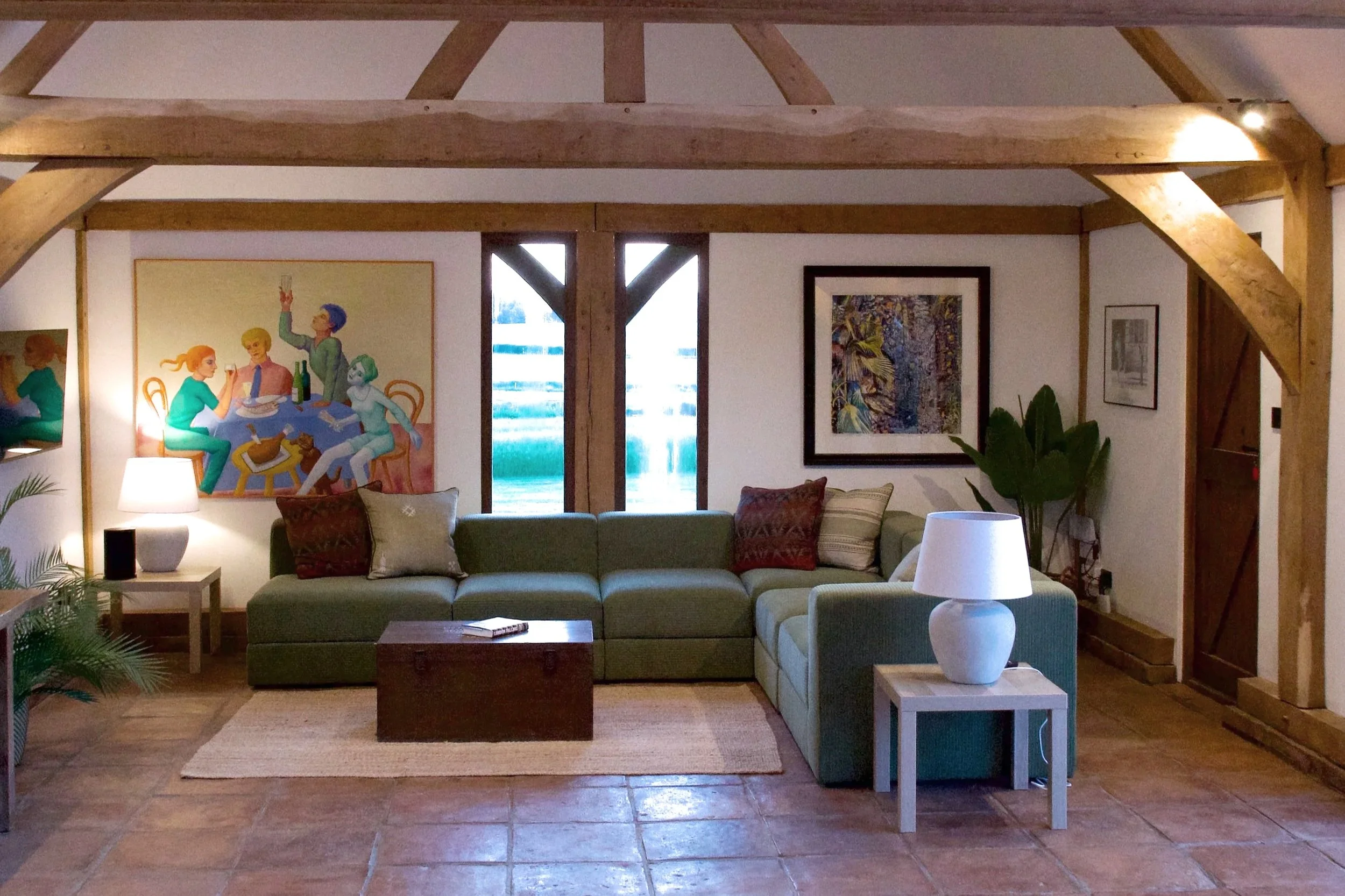 Living room with a green sectional couch, books, pillows, a white side table with a white lamp, wall art, and wooden beams in a rustic style.