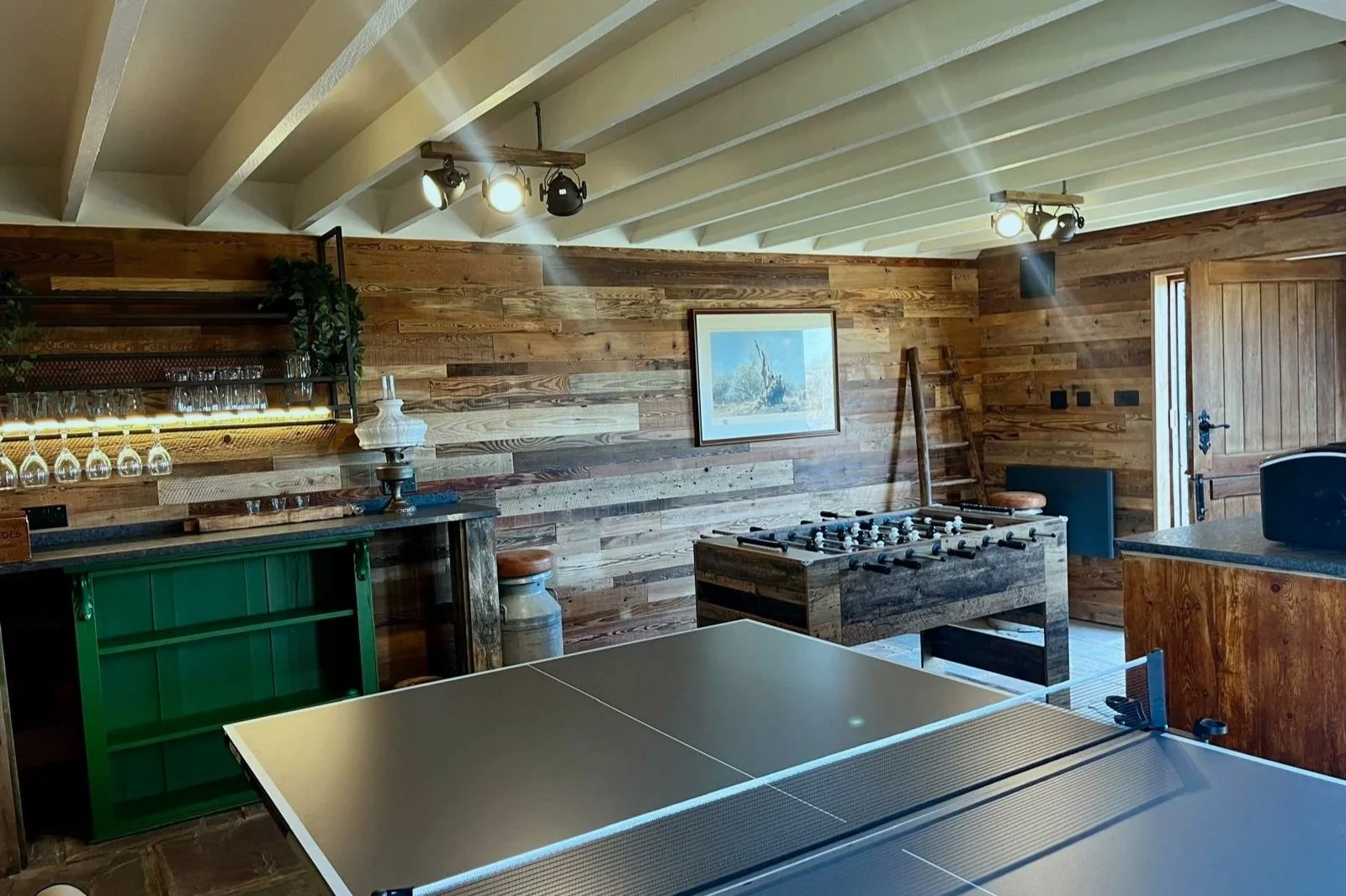 A rustic game room with wooden walls, a ping pong table in the foreground, a foosball table, a ladder, and a framed nautical painting on the wall. There are ceiling lights and glassware on shelves.