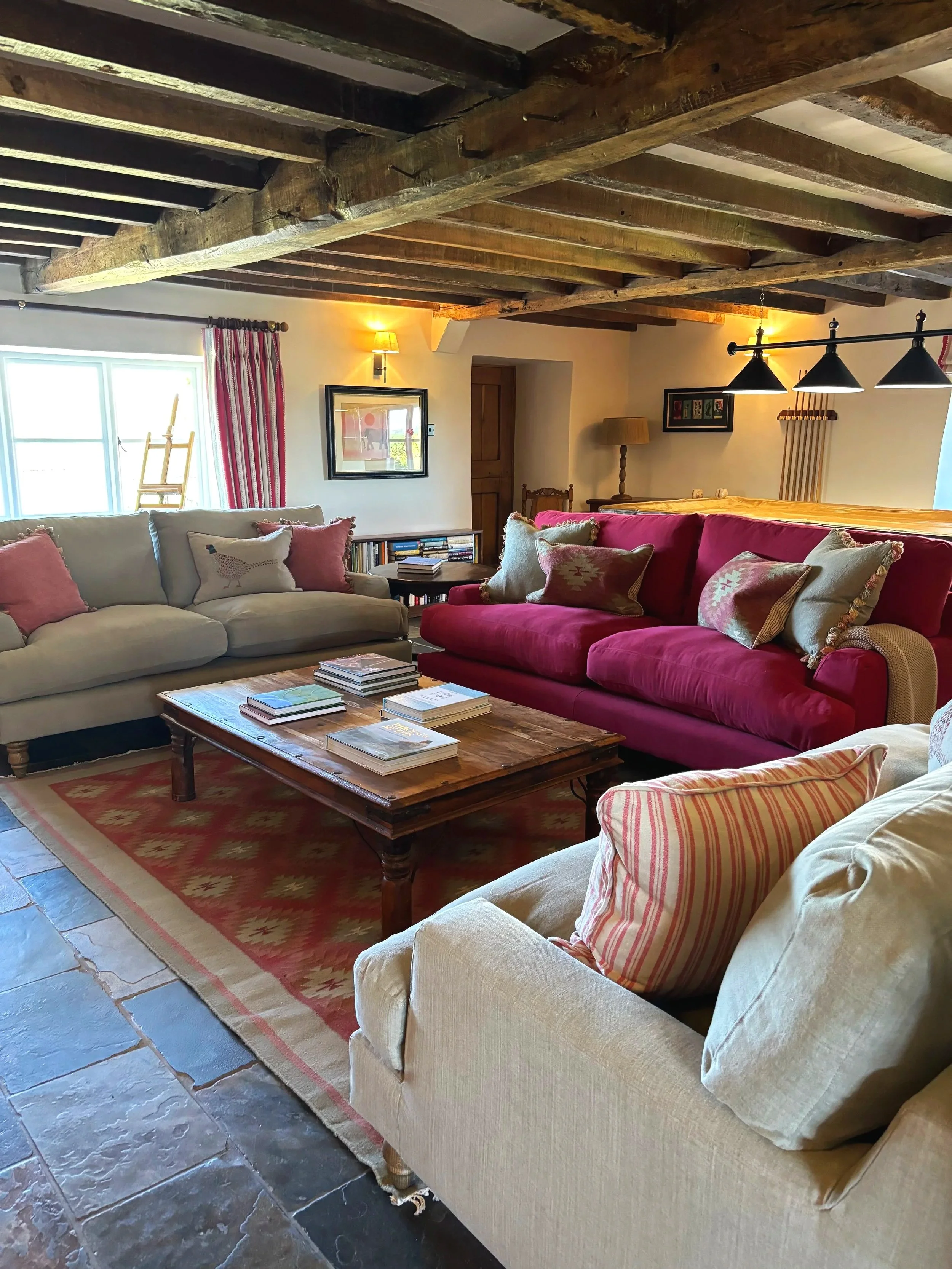 A cozy living room with two beige sofas, one with red and striped pillows and the other with pink pillows. A wooden coffee table with books is in front of the sofas. There is a window with pink and white striped curtains, a bookshelf, and wall art. T