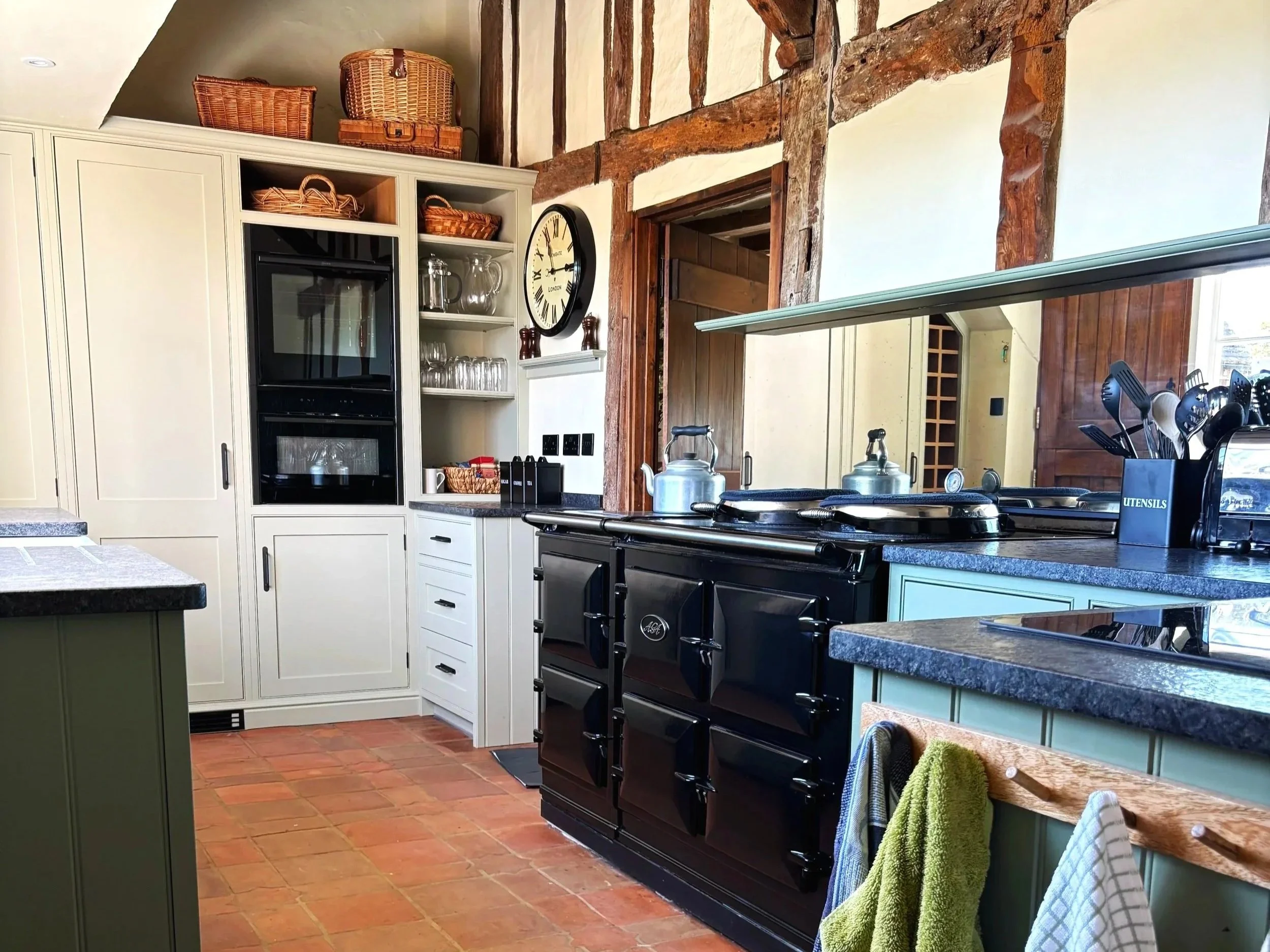 Kitchen with exposed wooden beams, black stove, white cabinetry, open shelves with glassware and baskets, clock on wall, and a mirror reflecting part of the room.