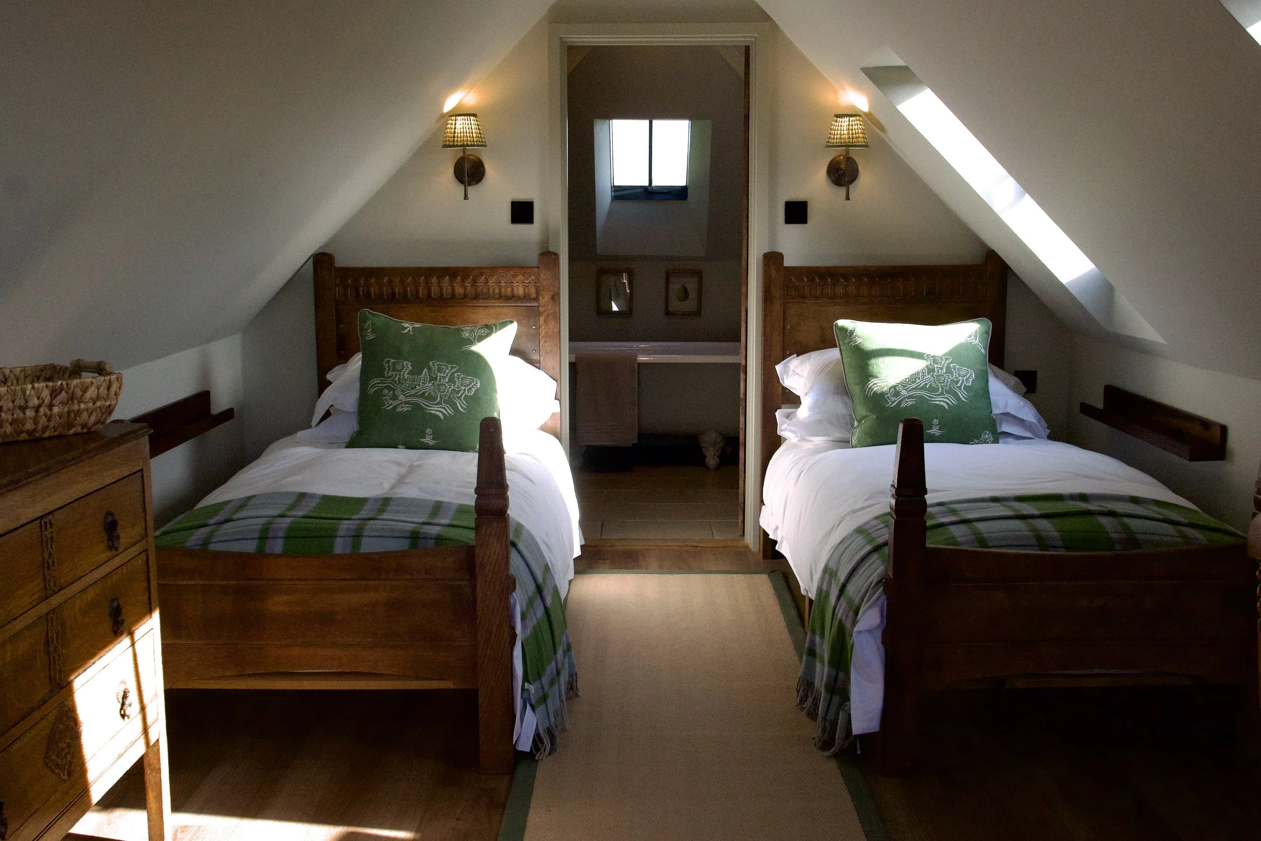 Two twin beds with wooden headboards and green pillows with white designs in a cozy attic bedroom, illuminated by natural light from a skylight and a window.