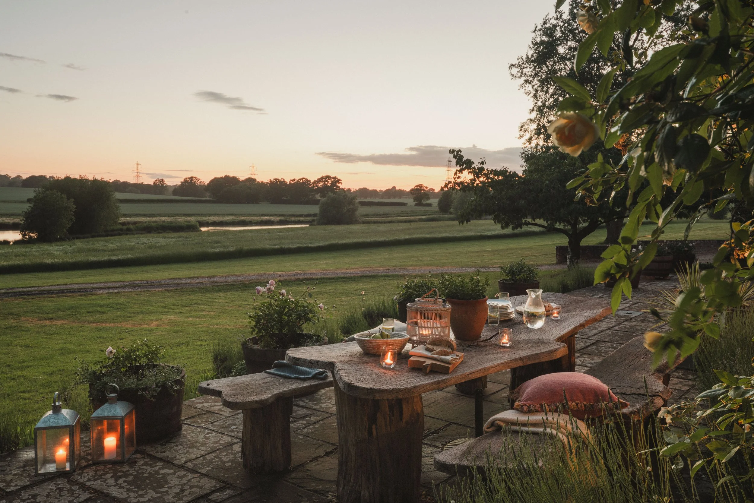 An outdoor patio scene at sunset with a rustic wooden table decorated with candles, bowls, and glasses, overlooking a grassy landscape with trees and power lines in the distance.