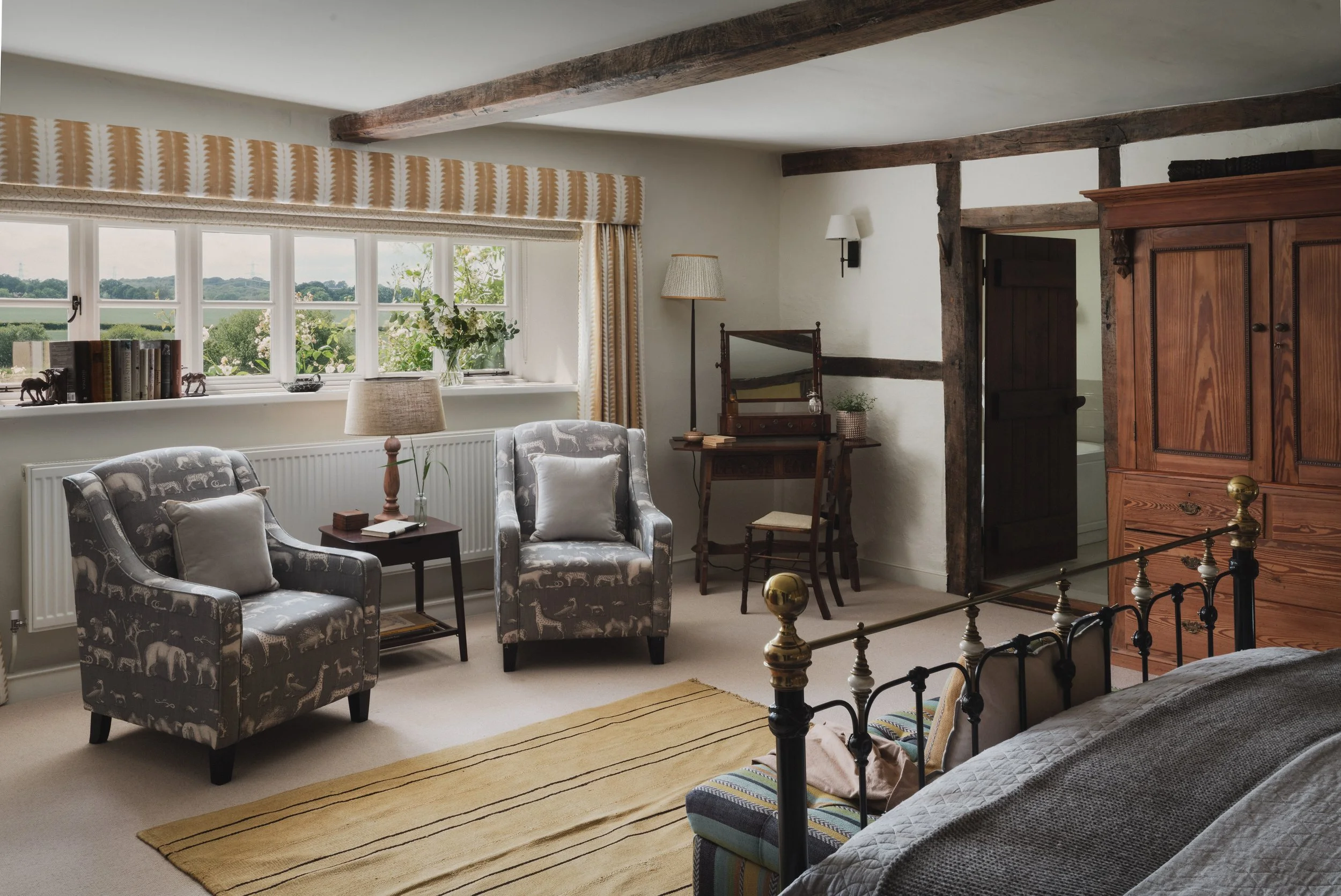 A cozy, rustic living room with a large window, patterned armchairs, a wooden armoire, a mirror, and decorative items like books and sculptures.