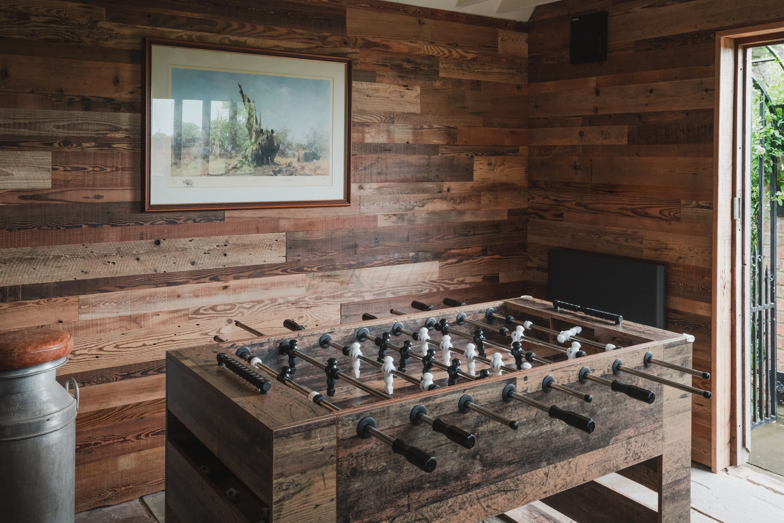 Indoor room with wooden walls, a framed picture of a bear outdoors on the wall, a foosball table in the center, and a metal milk canister on the left, near a stool. An open door reveals greenery outside.