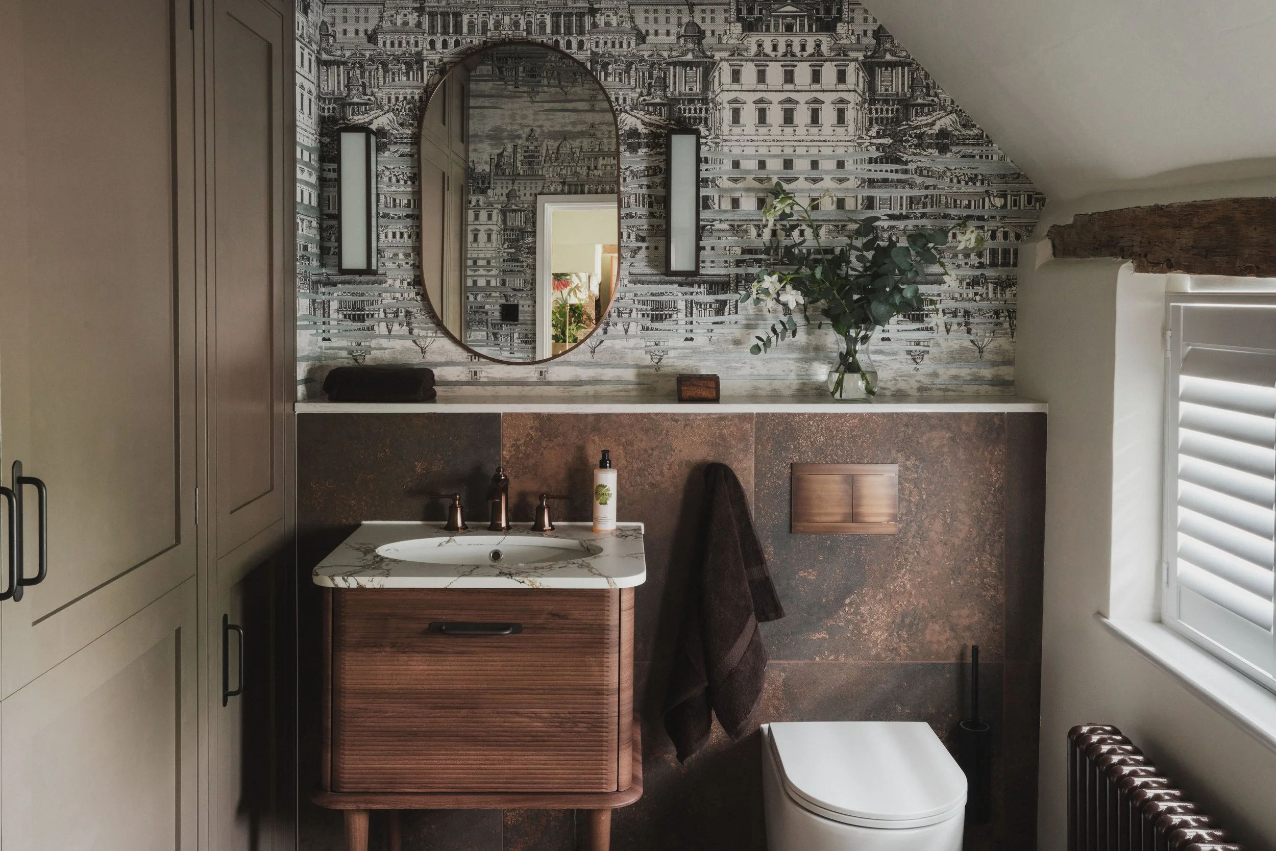 A modern bathroom with a wall-mounted mirror, decorative wallpaper with architectural illustrations, a small marble sink, a dark towel, a vase with green leafy stems, a brown wooden cabinet, and a window with plantation shutters.