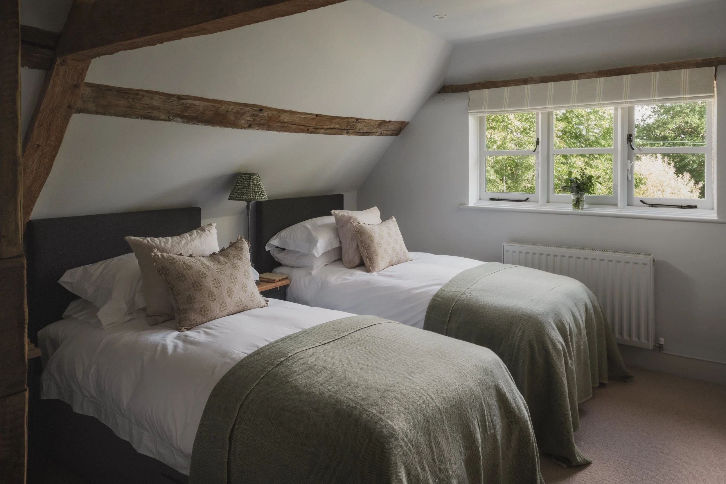 A cozy bedroom with two beds, each with white linens, beige and white pillows, and green bed runners. The room features a large window with a view of green trees outside, a radiator below the window, and a small nightstand with a green checkered lamp