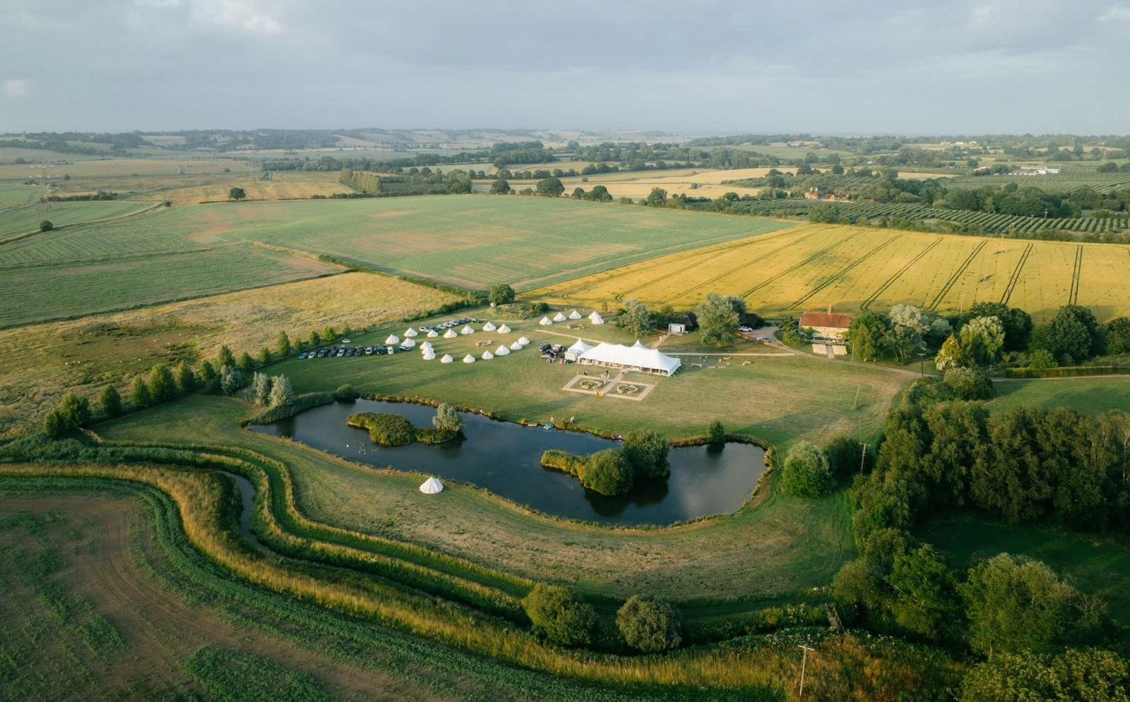 An aerial view of a large outdoor event with white tents set up near a pond, surrounded by green fields and farmland with a cloudy sky.