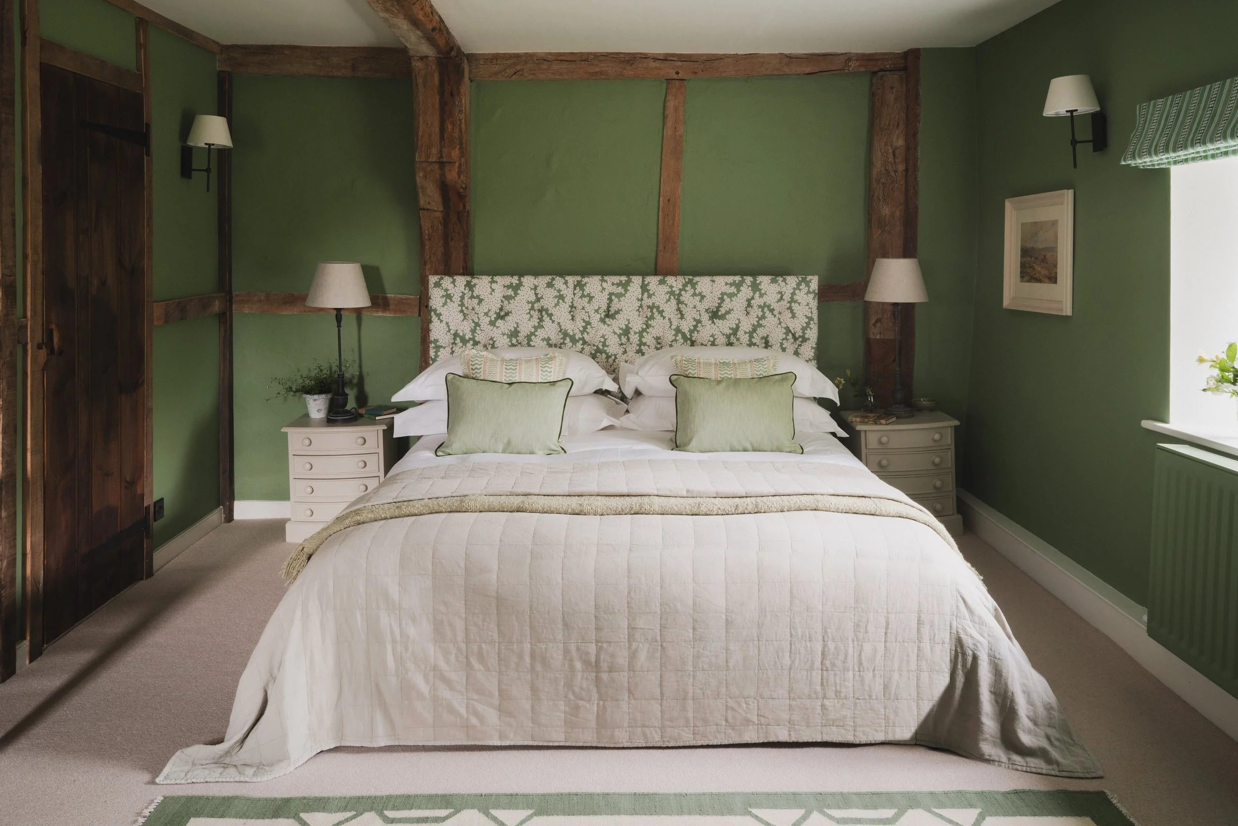 A bedroom with a bed that has a green and white floral headboard, white bedding with pillows, two nightstands with lamps, green walls, wooden beams, picture frame, and a window with a green and white striped curtain.