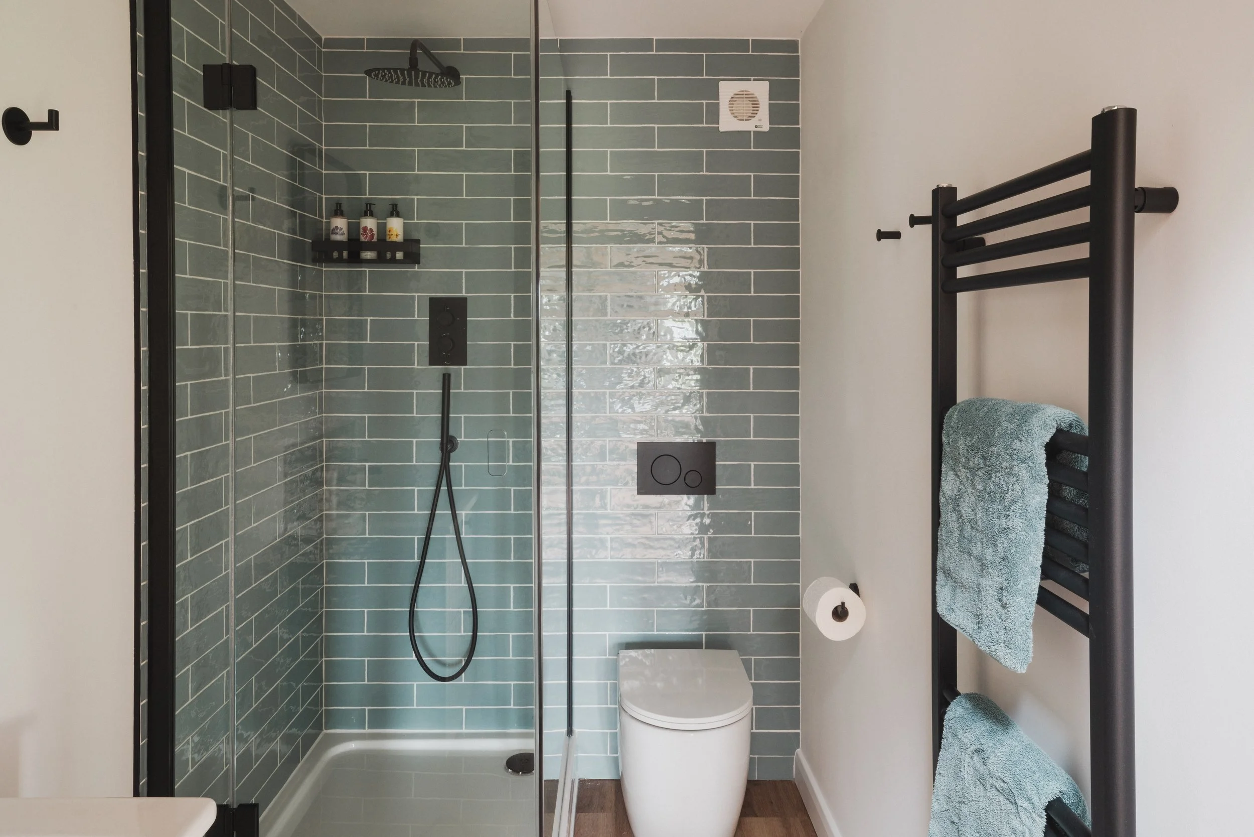 Modern bathroom with a walk-in shower featuring green subway tiles, a rainfall showerhead, and a black handheld shower. Adjacent to the shower is a white toilet and a black towel rack with a green towel. A small exhaust fan is on the white wall.