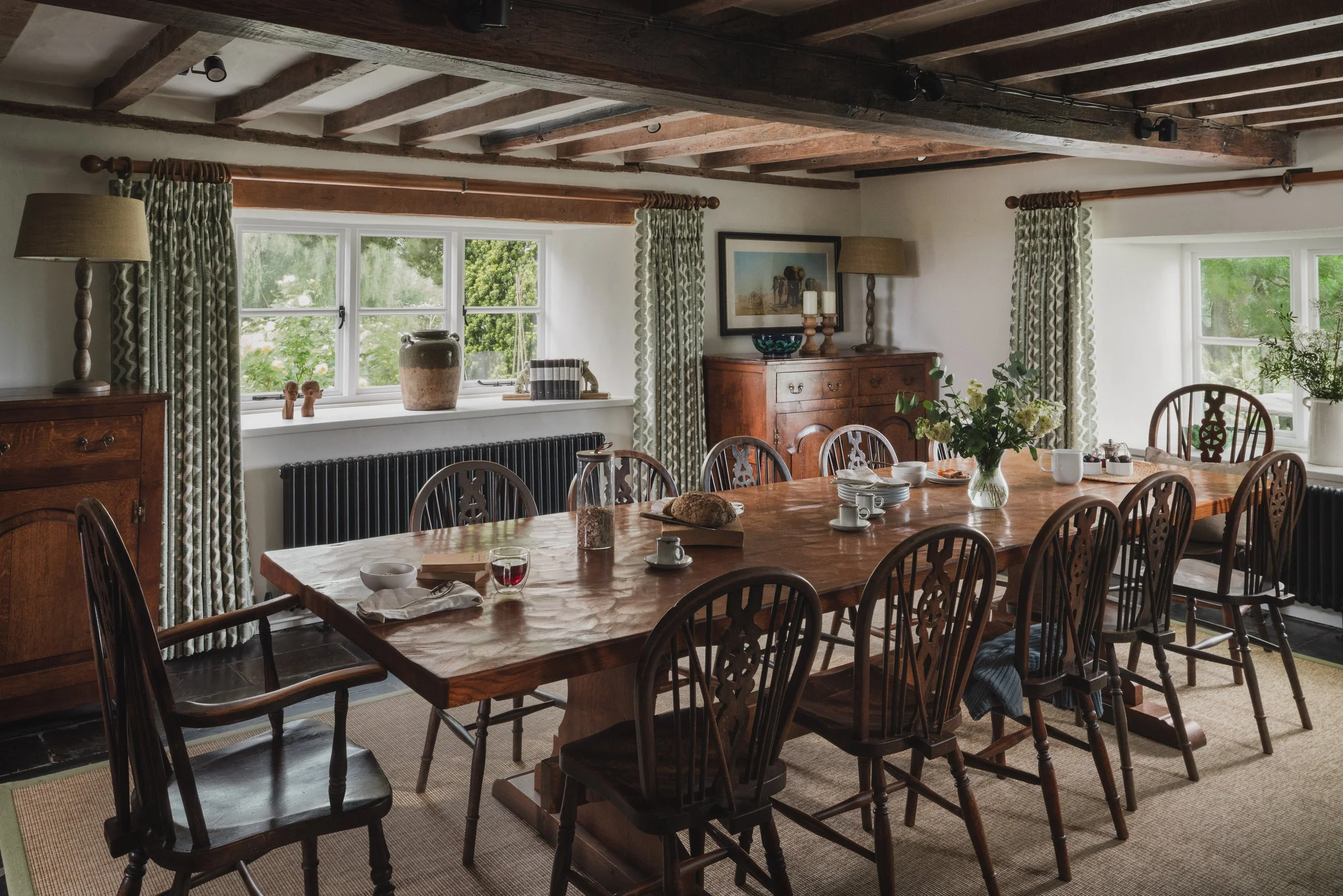 A dining room with a large wooden table, surrounded by wooden chairs, set with plates, cups, and a glass of red wine. The room has windows with patterned curtains, a wooden sideboard, and decorative items including vases, framed art, and a table lamp