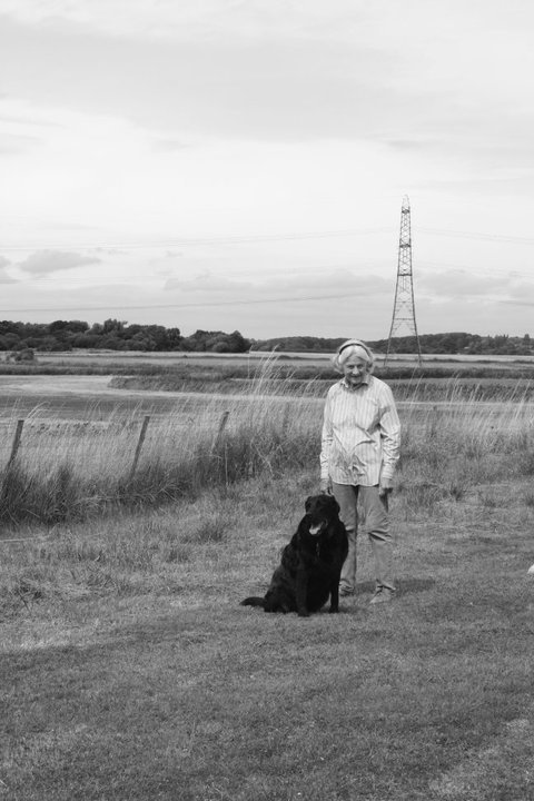 A woman standing in a grassy field with her black dog sitting beside her, with a power line tower and open landscape in the background.
