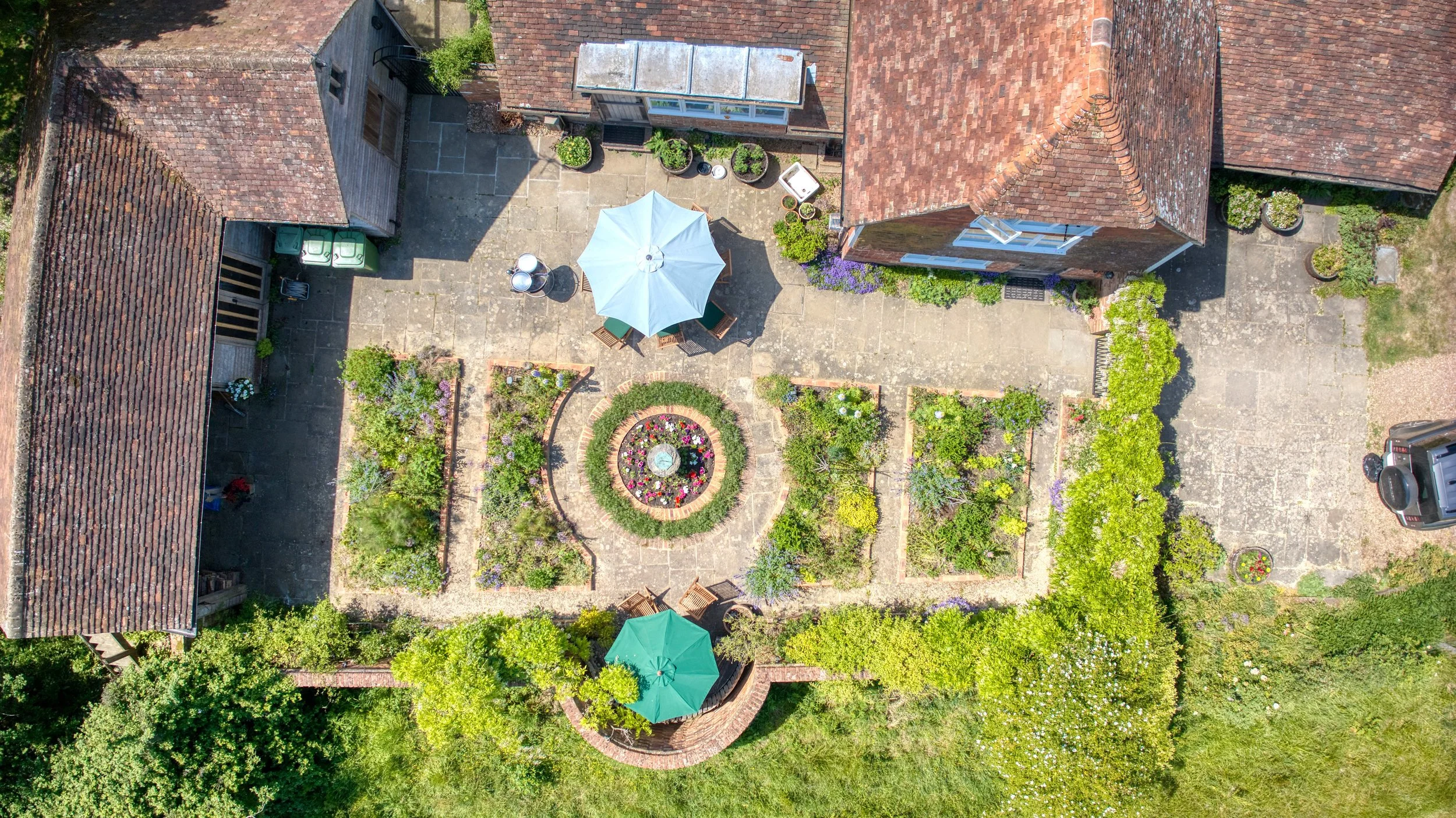 An aerial view of a backyard garden with a patio, a white umbrella, outdoor furniture, potted plants, flower beds, and adjacent rooftops.