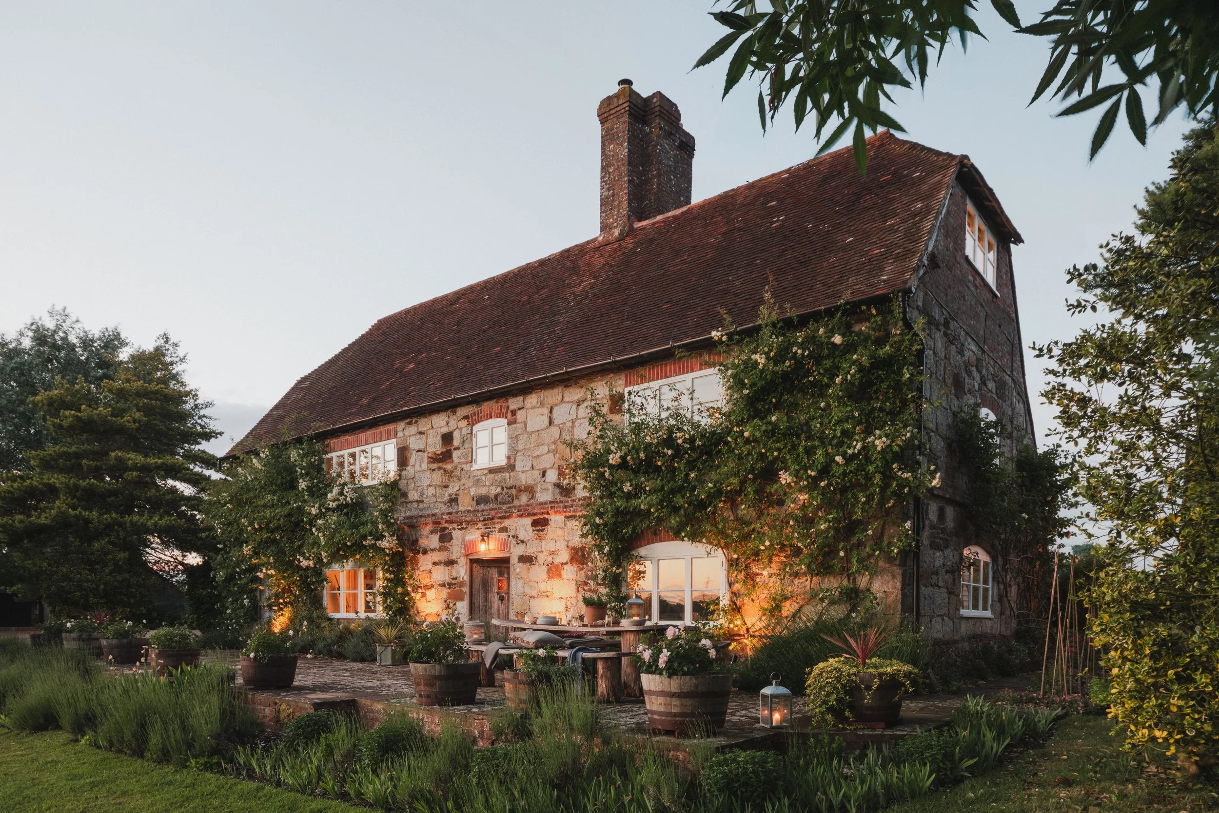 A rustic brick and stone house with a steep red-tiled roof, chimney, and white-framed windows, surrounded by green trees and plants, with outdoor string lights and potted plants illuminating the yard during dusk.