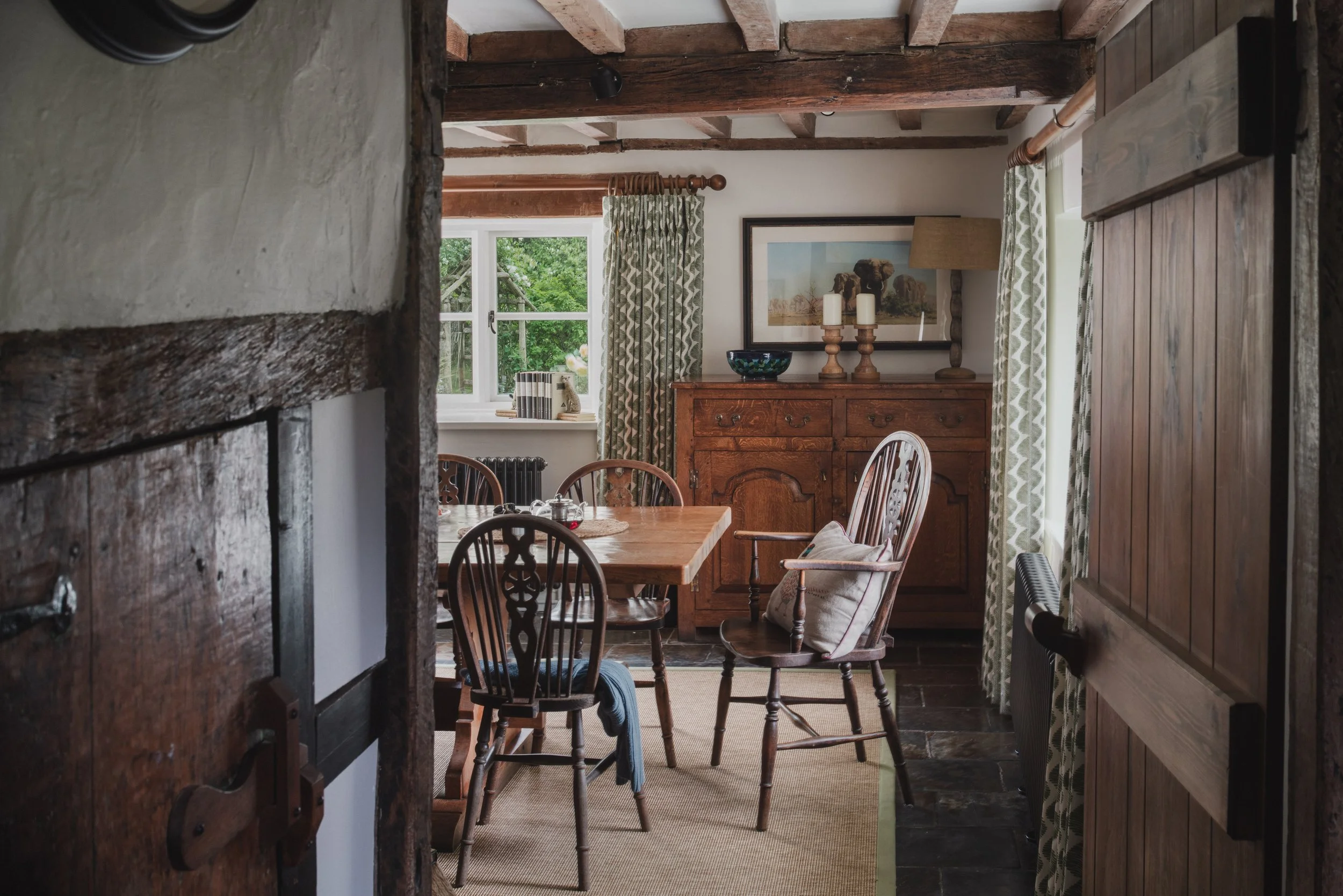 Interior view of a cozy dining room with wooden beams, a wooden sideboard, framed artwork, and a window with patterned curtains, seen from an open doorway.
