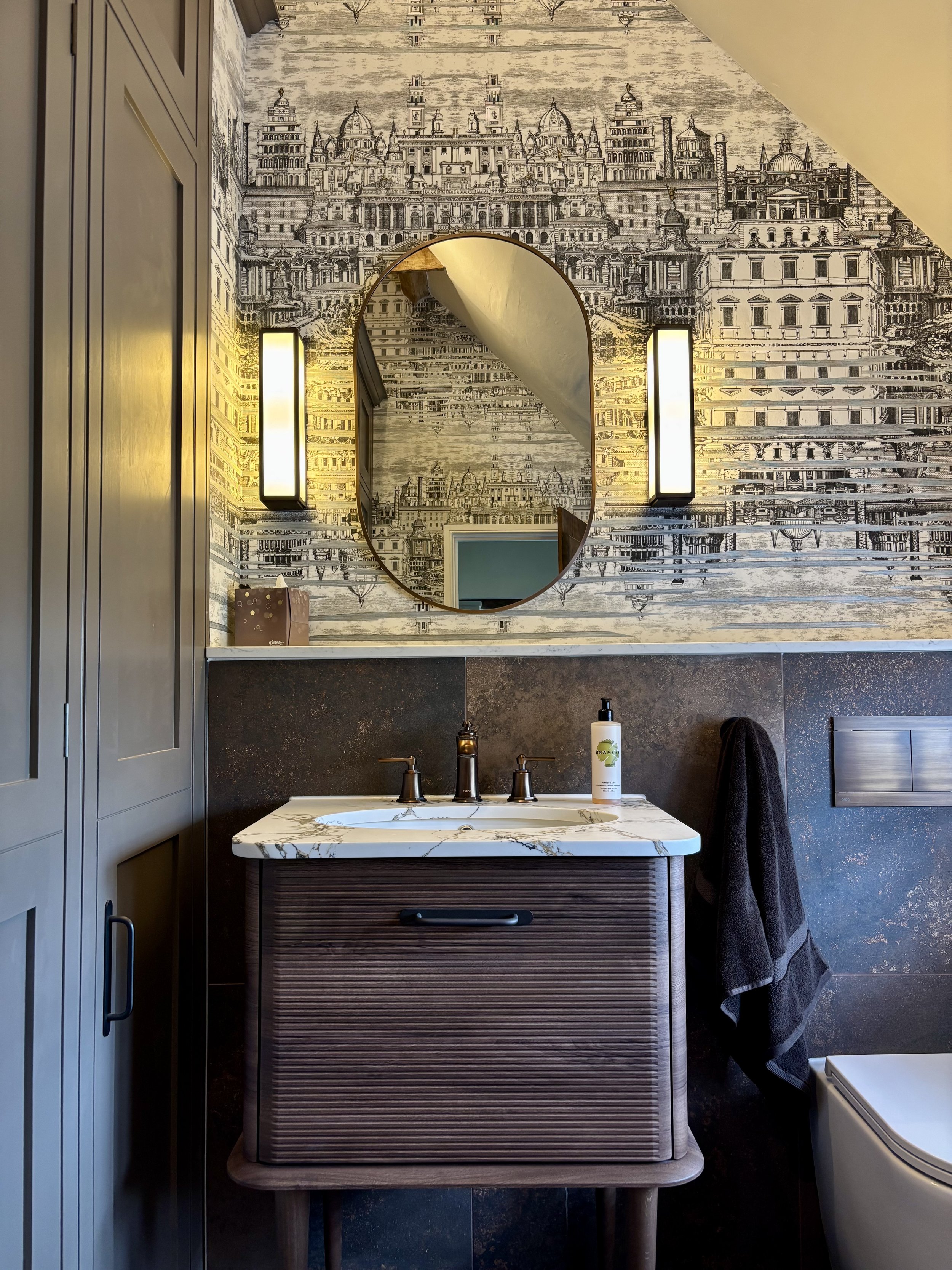 A bathroom corner with a wall decorated with black and white cityscape mural, a mirror, two vertical light fixtures, a sink with a marble countertop, and a brown wooden cabinet with a handle. A bottle of hand soap and a black towel are on the counter