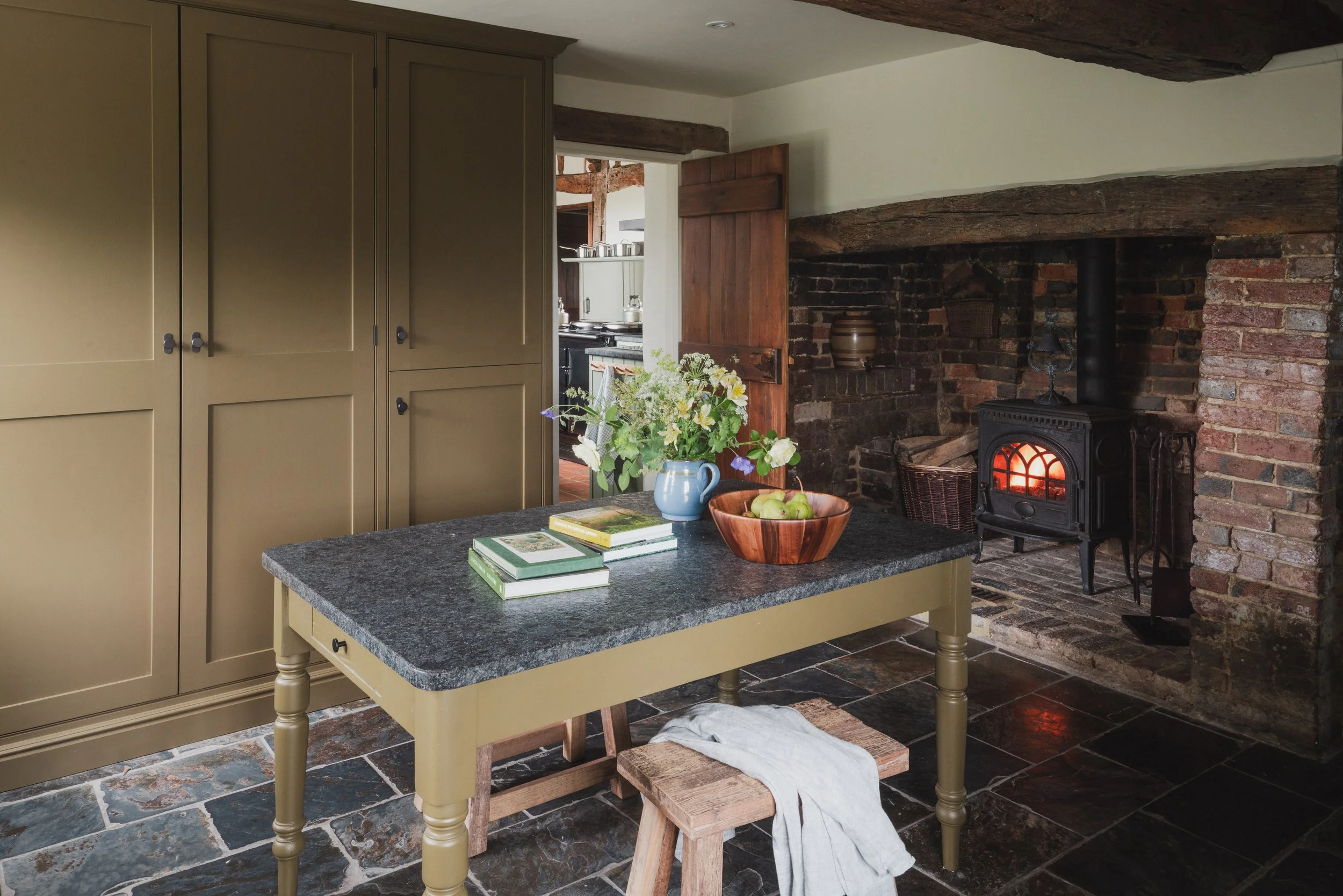 A cozy kitchen with a rustic brick fireplace and a wood stove. A green kitchen island with a dark granite countertop features a blue pitcher with fresh flowers, a wooden bowl of apples, and several books. Light-colored cabinets line the wall, and the