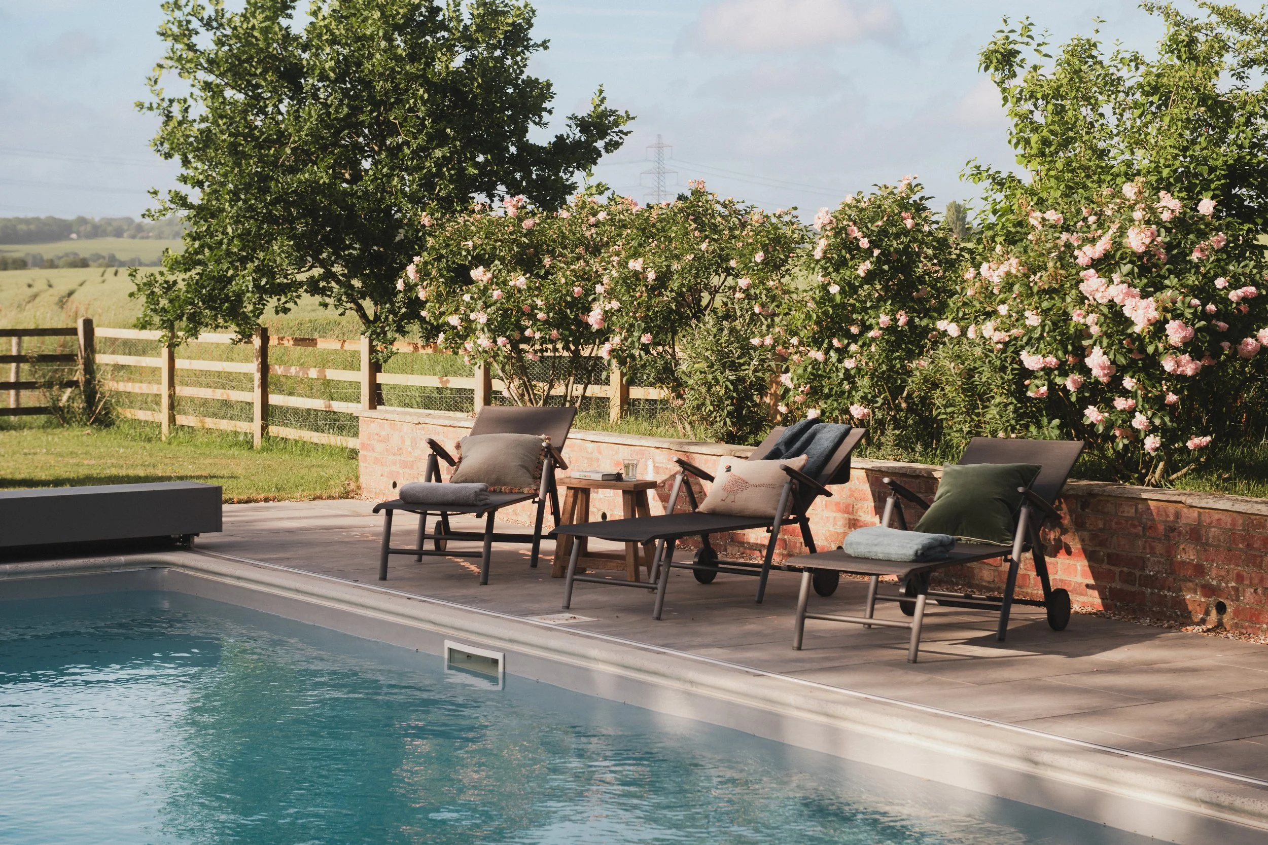 A backyard swimming pool area with lounge chairs and pillows, surrounded by a brick wall and a wooden fence, with trees and pink flowering bushes in the background.