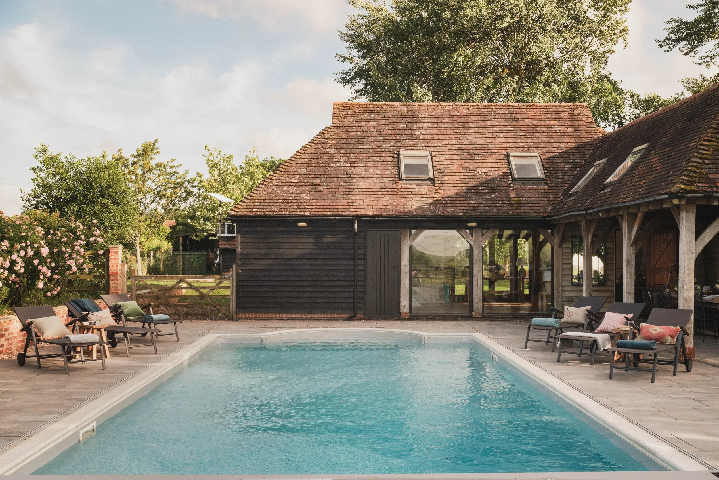 A backyard with a swimming pool surrounded by lounge chairs and an outdoor seating area with cushions. In the background, there's a rustic house with a sloped roof and skylights, and green trees under a partly cloudy sky.