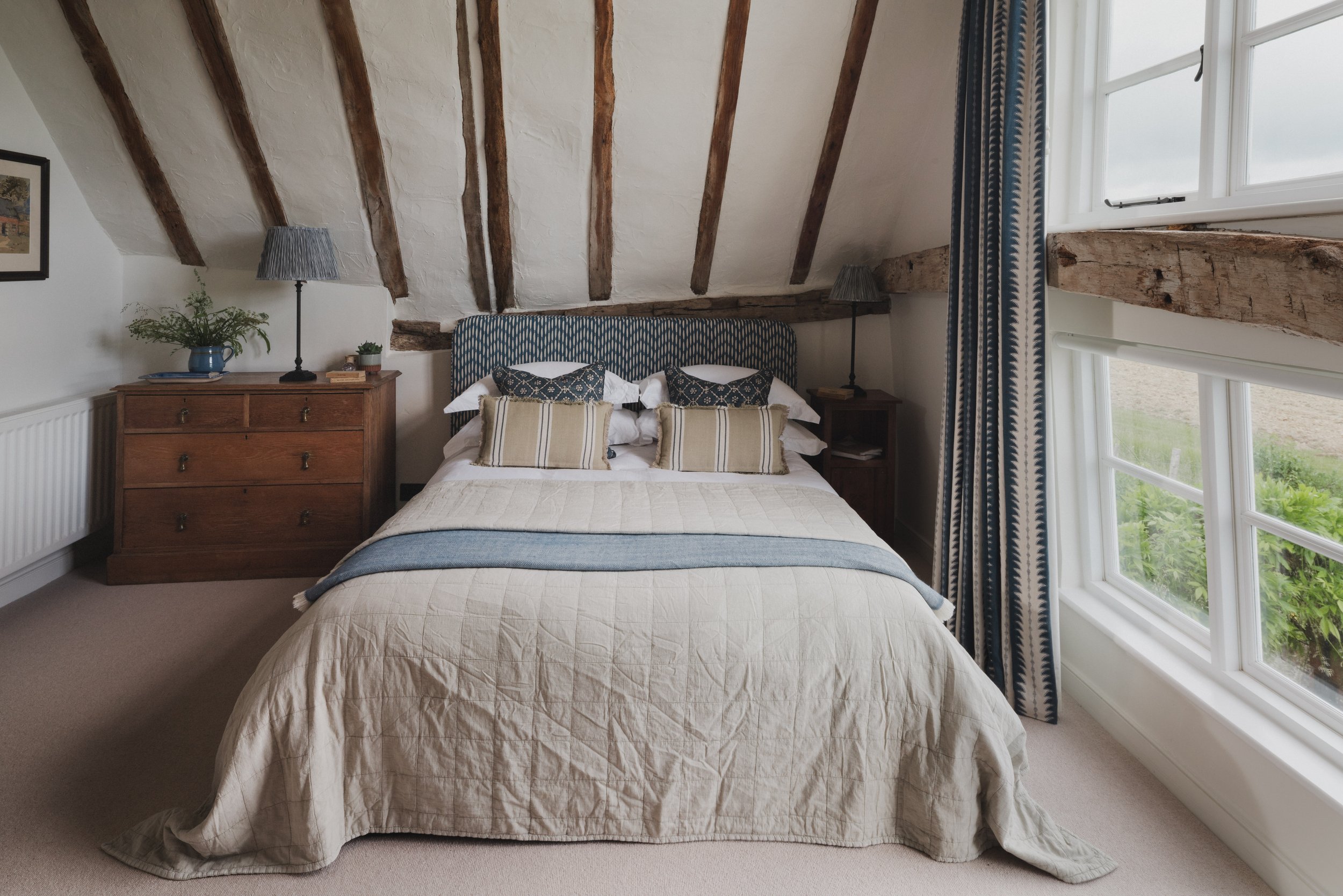 A cozy bedroom with a sloped ceiling, a double bed with beige quilt, multiple decorative pillows, wooden side tables with lamps, a dresser, a plant, and a large window with patterned curtains showing lush greenery outside.