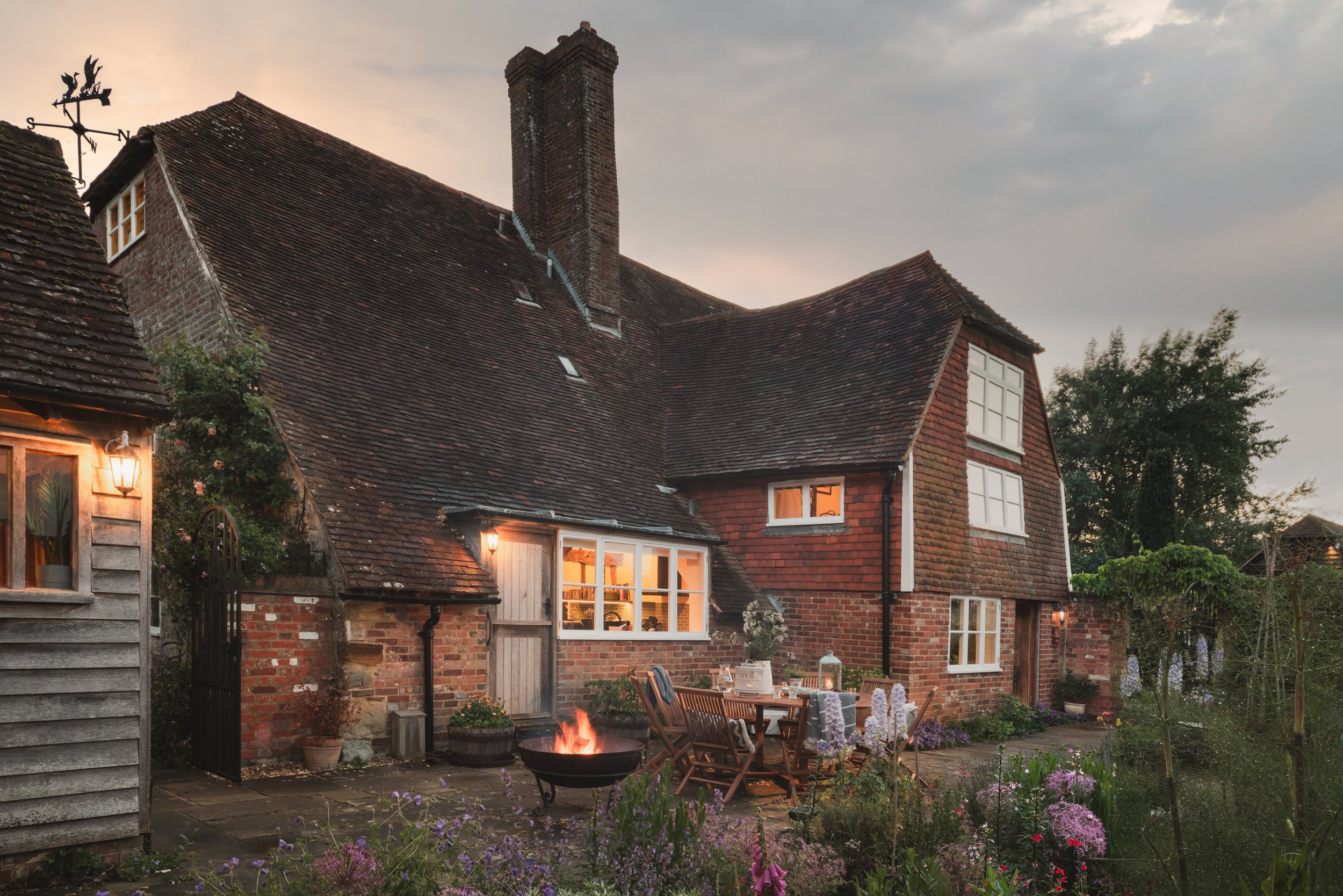 A cozy brick house with a steep roof, large windows with warm interior lighting, and a backyard patio with a fire pit, outdoor table, chairs, and blooming flowers during dusk.