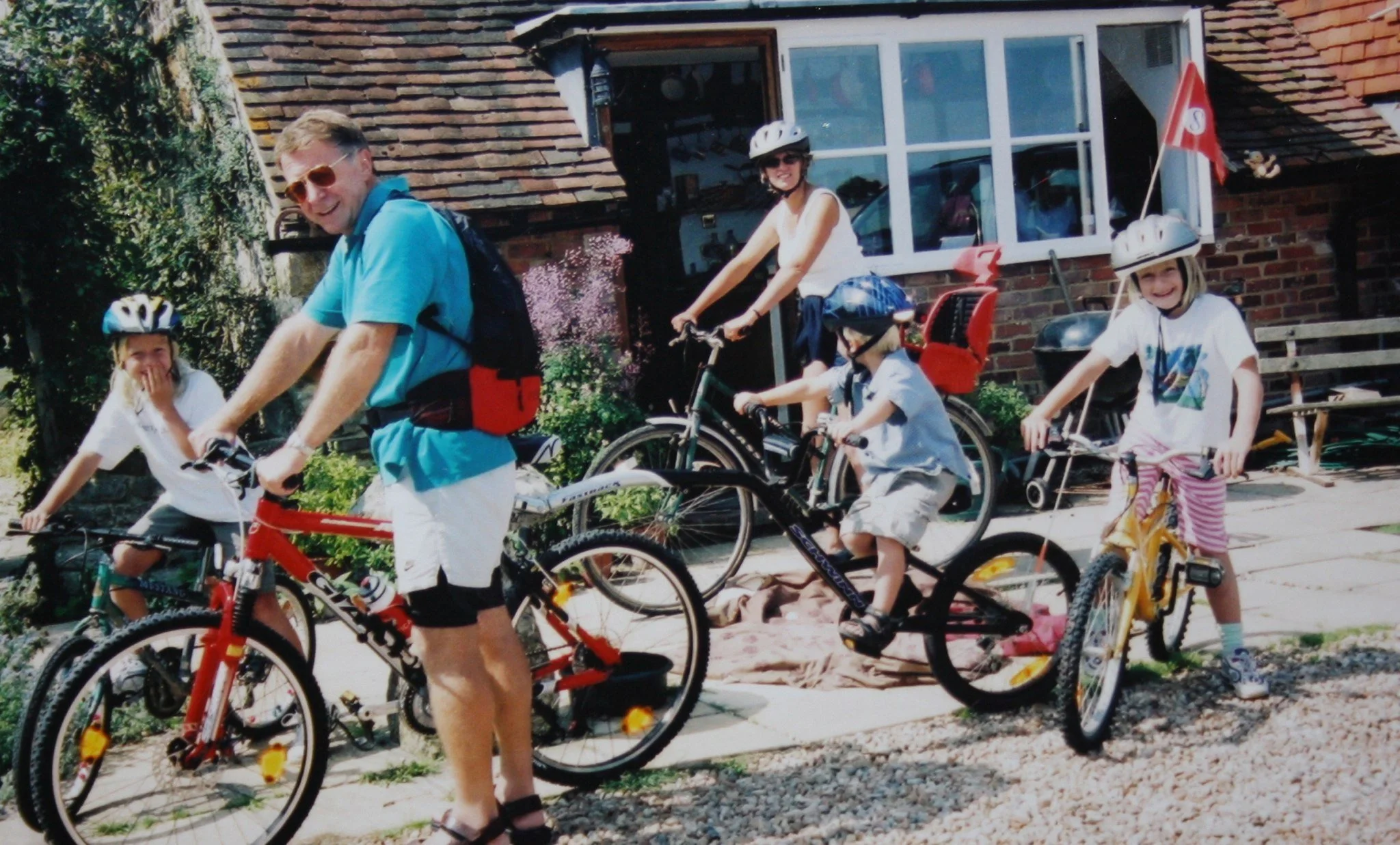A man and four children riding bicycles in front of a brick house with a window. All are wearing helmets, and the children are smiling.