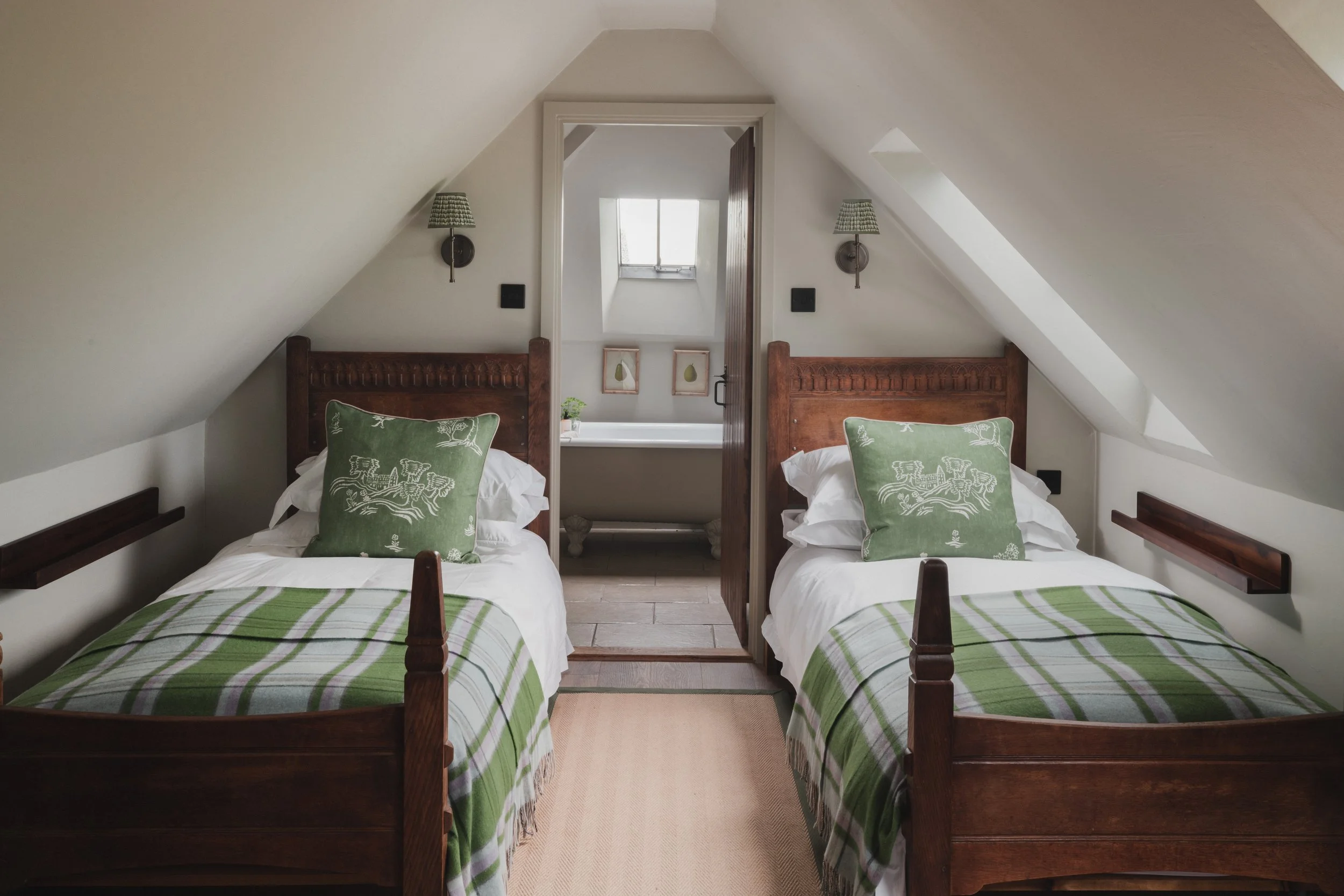 Two twin beds with green and white plaid bedding and decorative green pillows in a cozy attic bedroom with sloped ceilings, wall-mounted lamps, a small window, and framed art pieces.