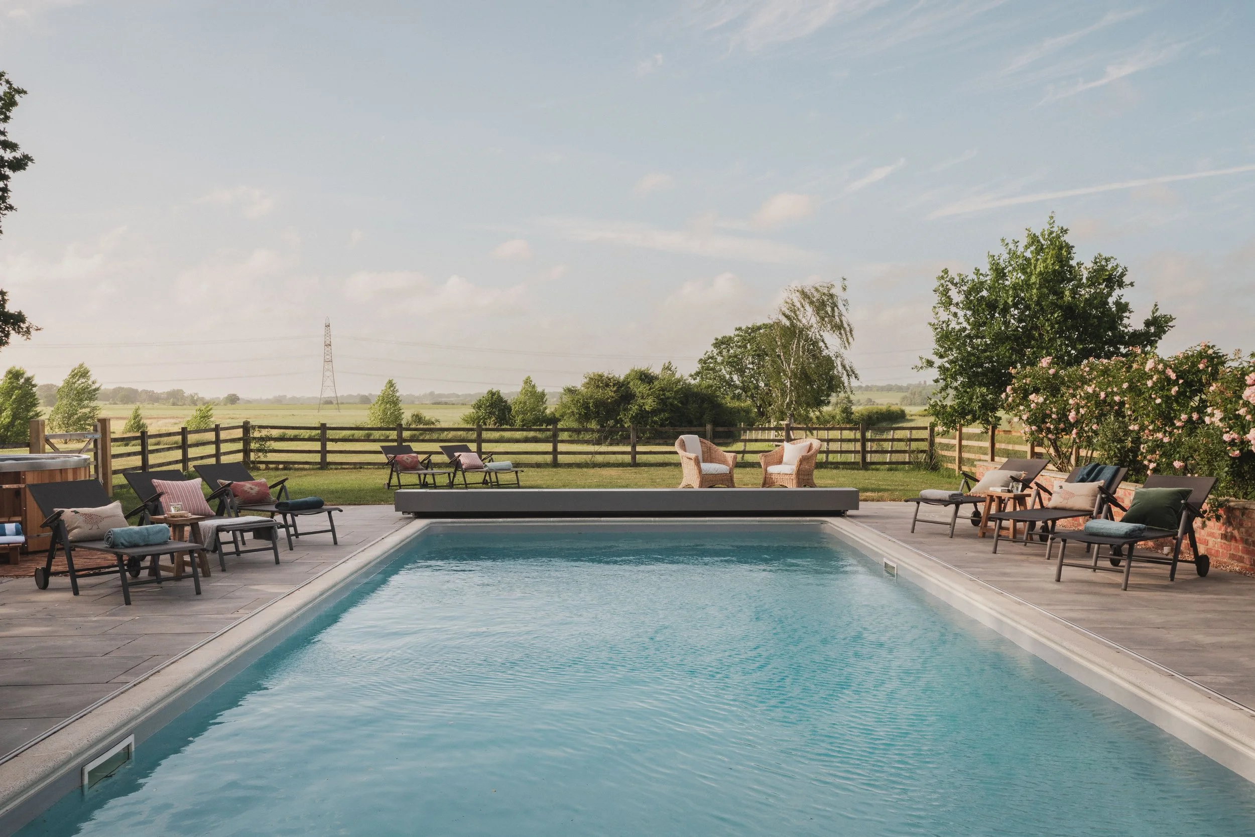 An outdoor swimming pool with lounge chairs and cushions on a patio, surrounded by a wooden fence and green trees under a partly cloudy sky.