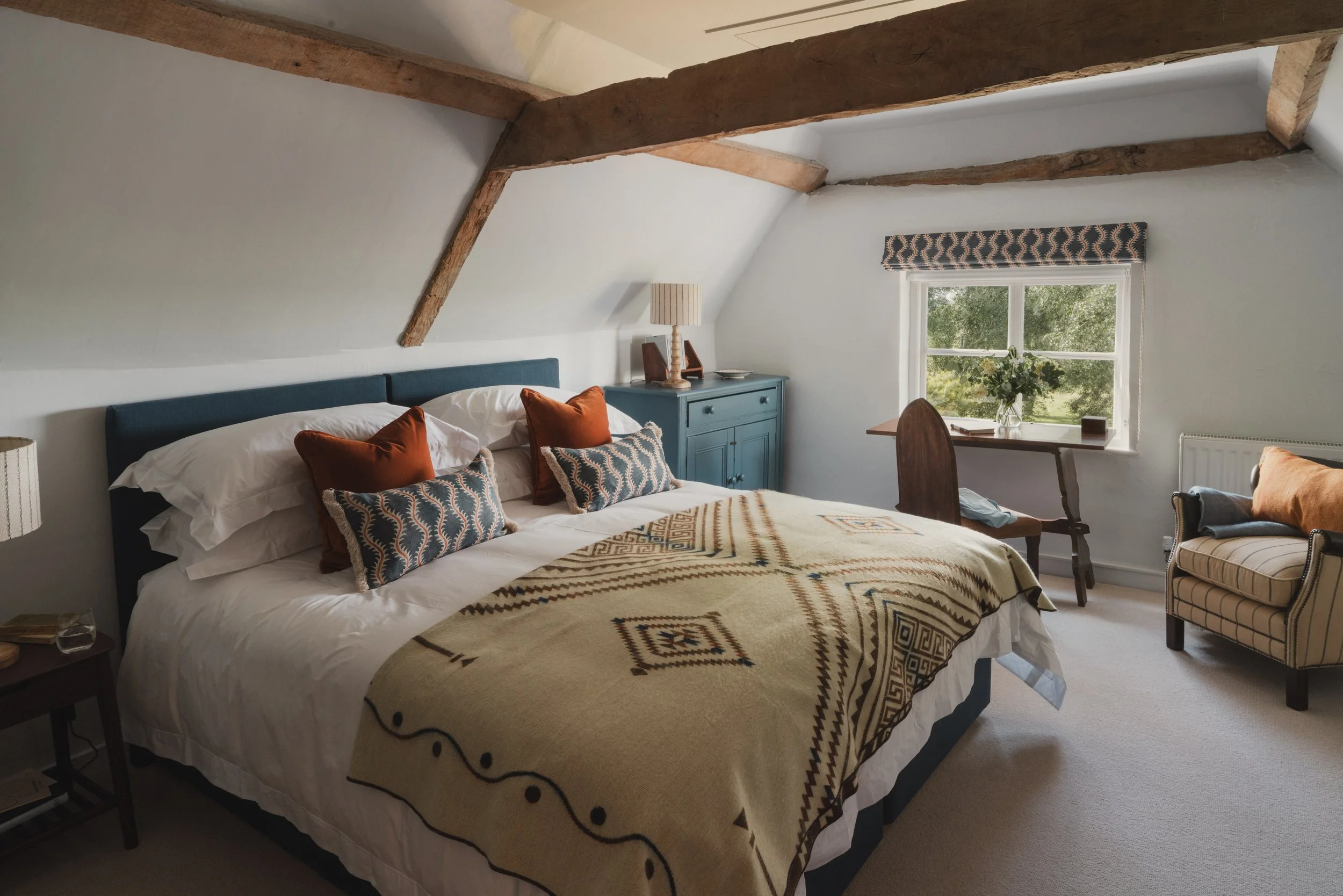 A cozy bedroom featuring a bed with white sheets, decorative pillows, and a patterned beige blanket. There is a blue bedside table with a lamp, a small chair, and a window with a view of greenery outside. The ceiling has exposed wooden beams.