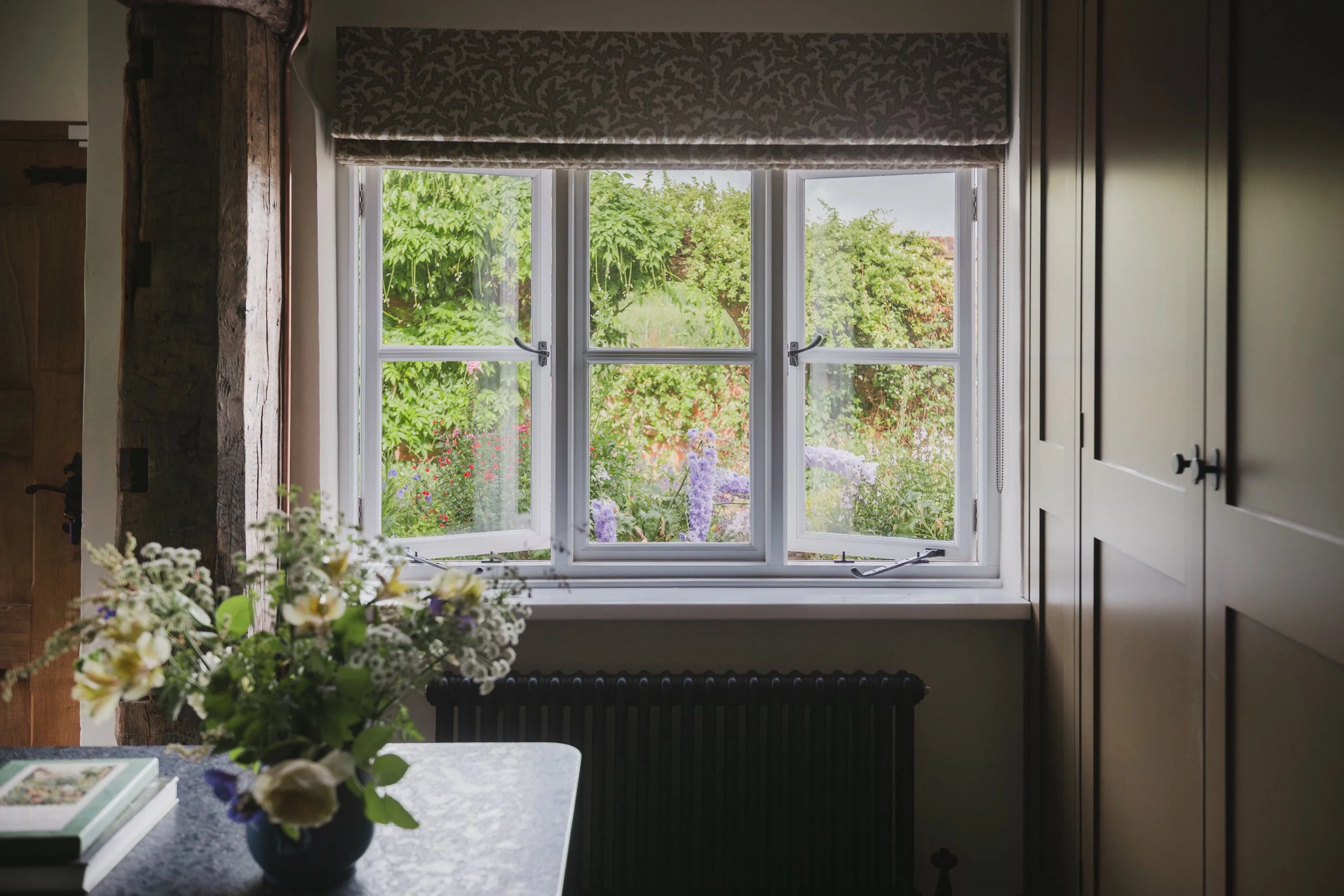 A kitchen window with white frame open, revealing a garden with green trees and colorful flowers outside. The kitchen has dark cabinets on the right and a wooden beam on the left. A vase of mixed flowers is on a table in the foreground.