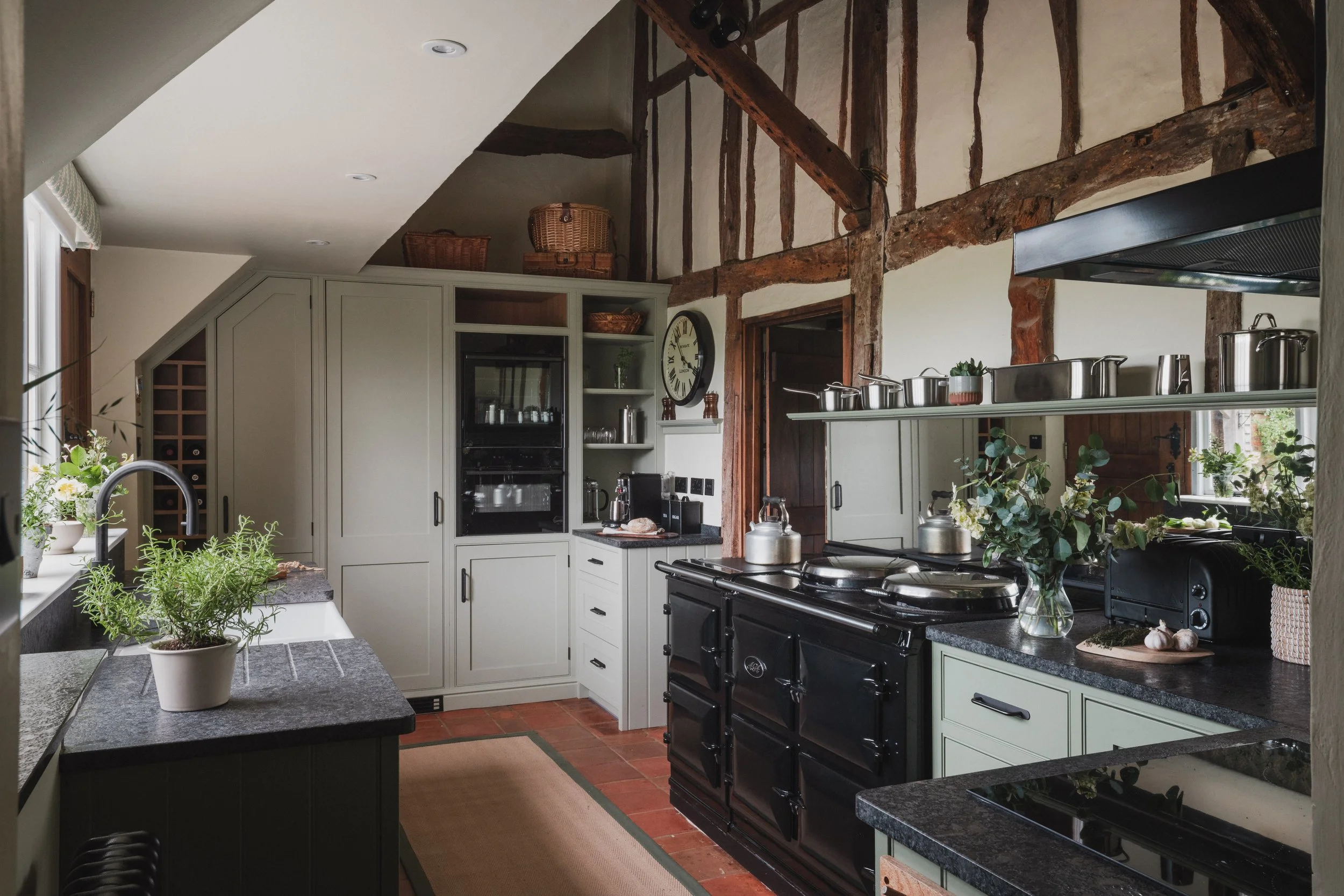 A kitchen with a rustic style, featuring exposed wooden beams, white cabinetry, black stove, and various pots on the stovetop. There are green plants on the counter and a wall clock on the wall. Natural light comes through a window.