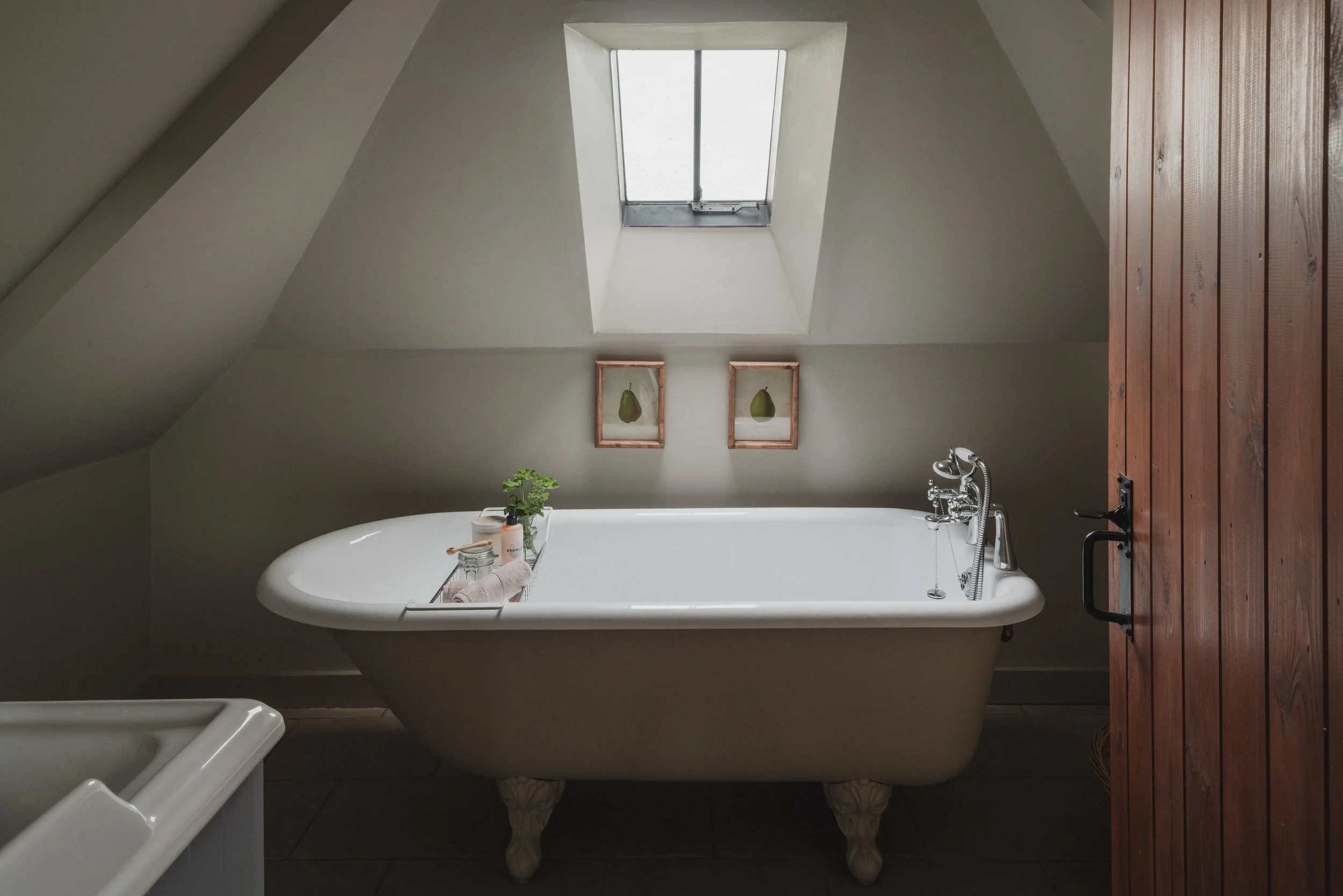 A bathroom with a vintage clawfoot bathtub under a skylight window. The tub has a plant, toiletries, and a towel on a rack. There are two framed pear prints on the wall and a wooden door on the right side.
