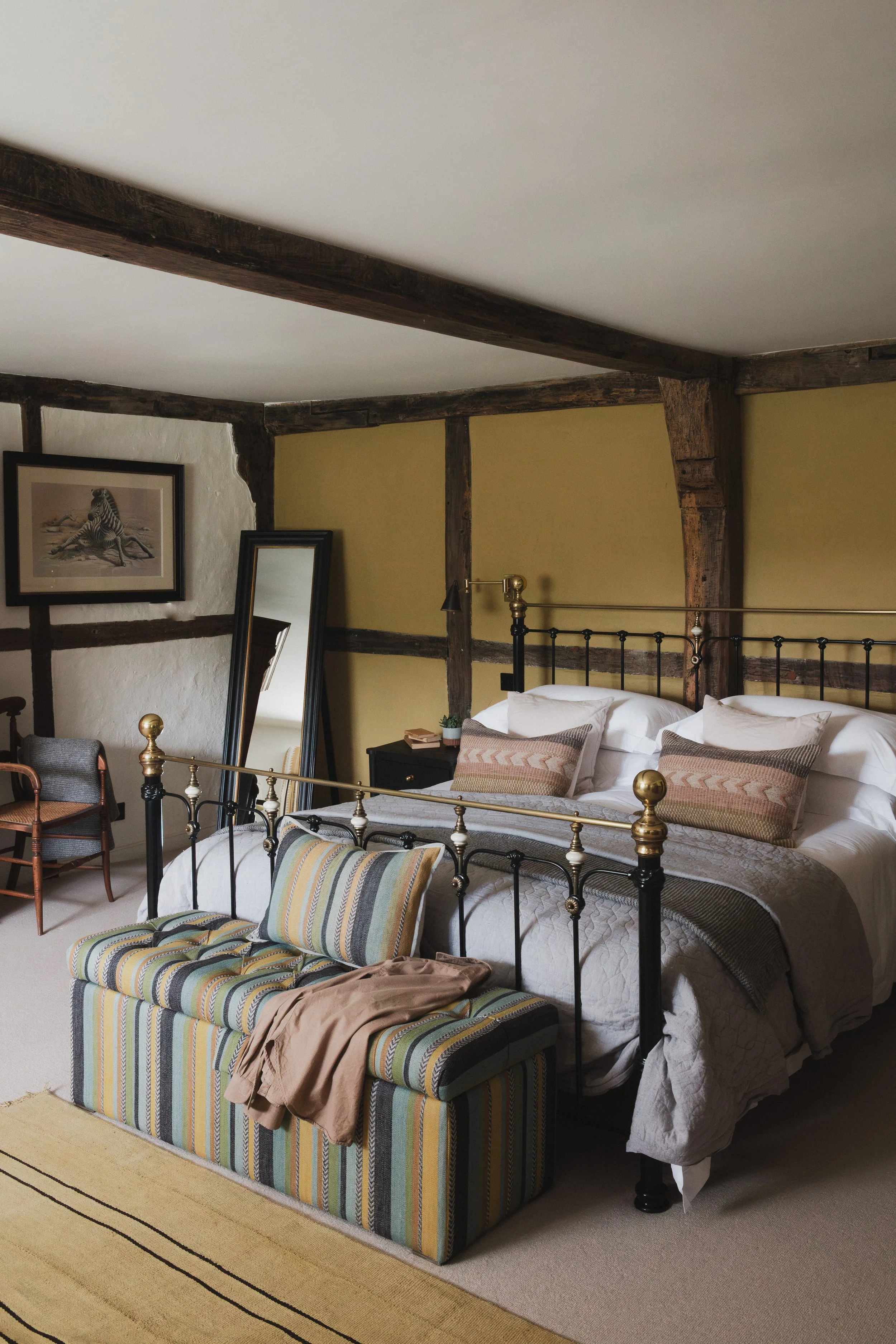 A rustic bedroom with exposed wooden beams on the ceiling and walls, a metal bed with white bedding and decorative pillows, an upholstered bench at the foot of the bed with a beige jacket and striped pillows, a full-length mirror leaning against the 