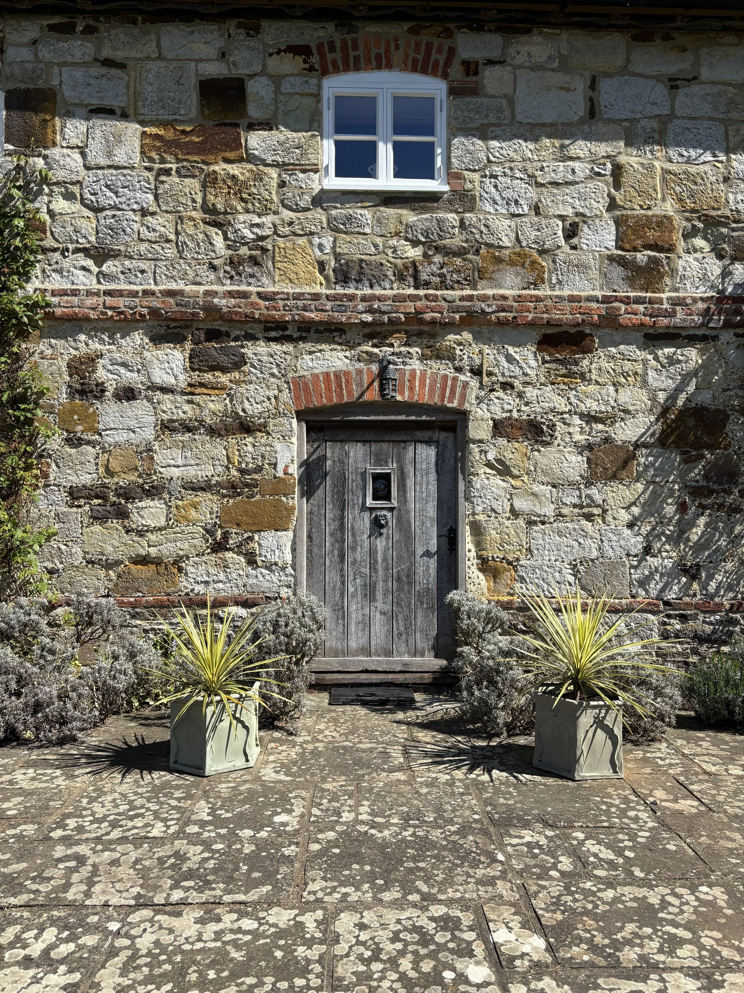 A rustic stone wall with a window and a wooden door, surrounded by potted plants and a stone patio.