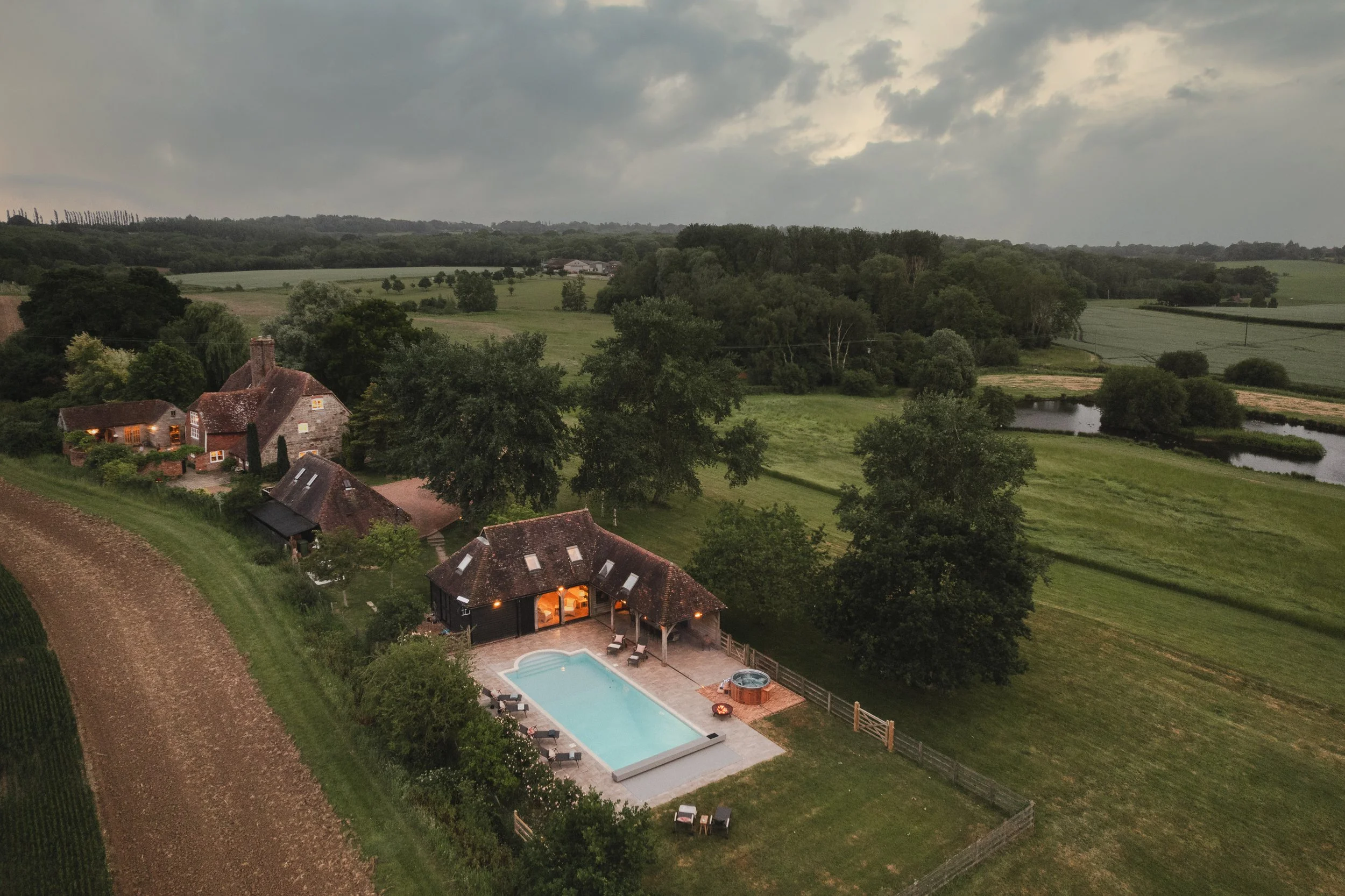 Aerial view of a countryside estate at dusk with a house, a swimming pool, green fields, trees, hills, and an overcast sky.
