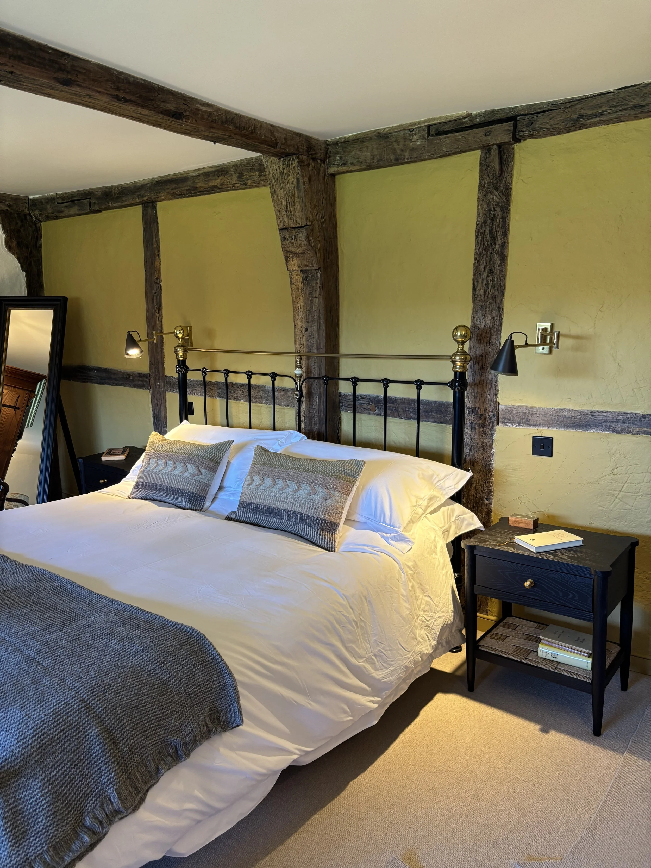 A cozy bedroom with a white bed featuring pillows, a black metal headboard with brass details, a black nightstand with books, and two black wall-mounted reading lamps attached to a rustic wooden beam ceiling painted yellow.