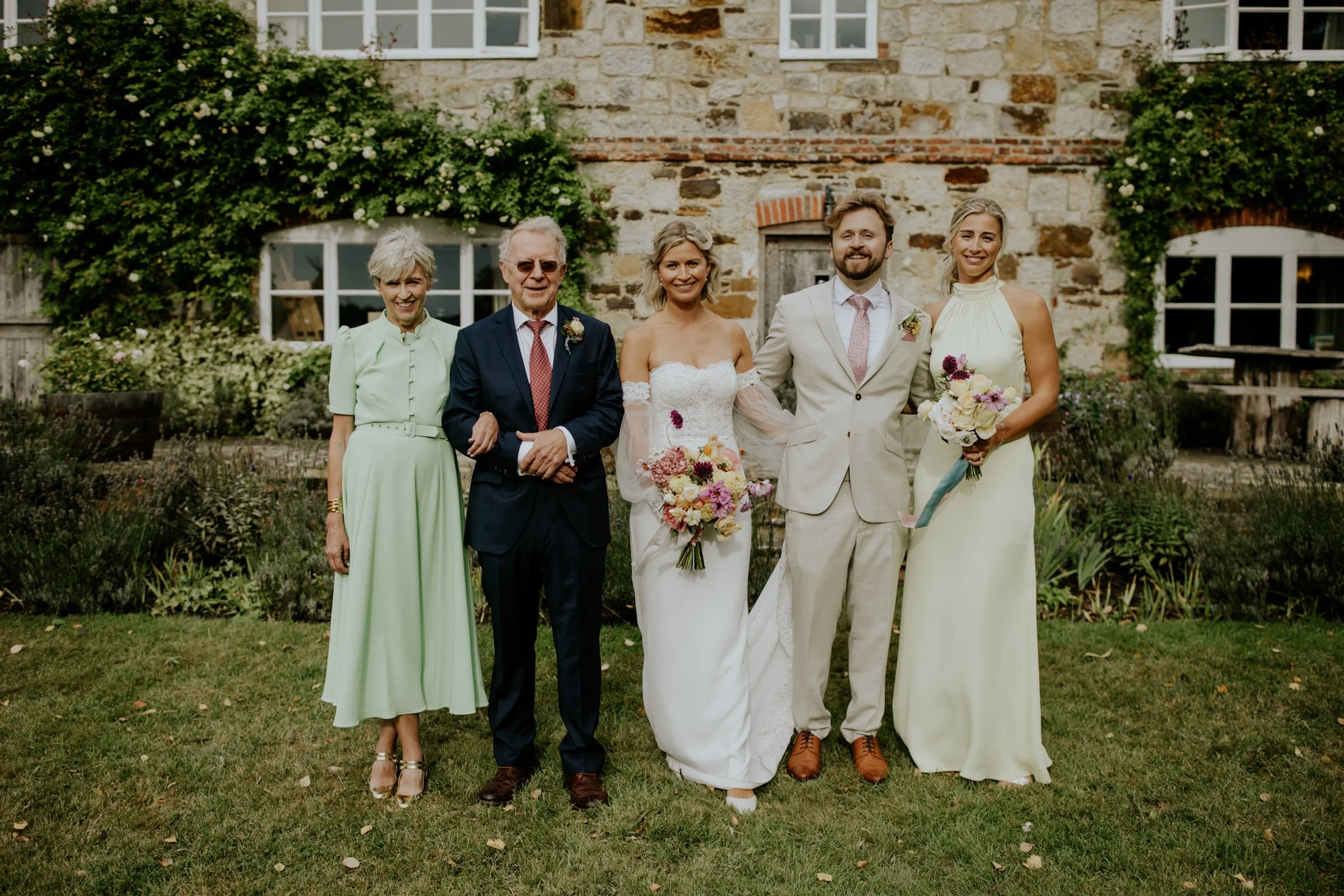 Group of five people standing in a garden in front of a stone house, dressed in wedding attire and smiling.