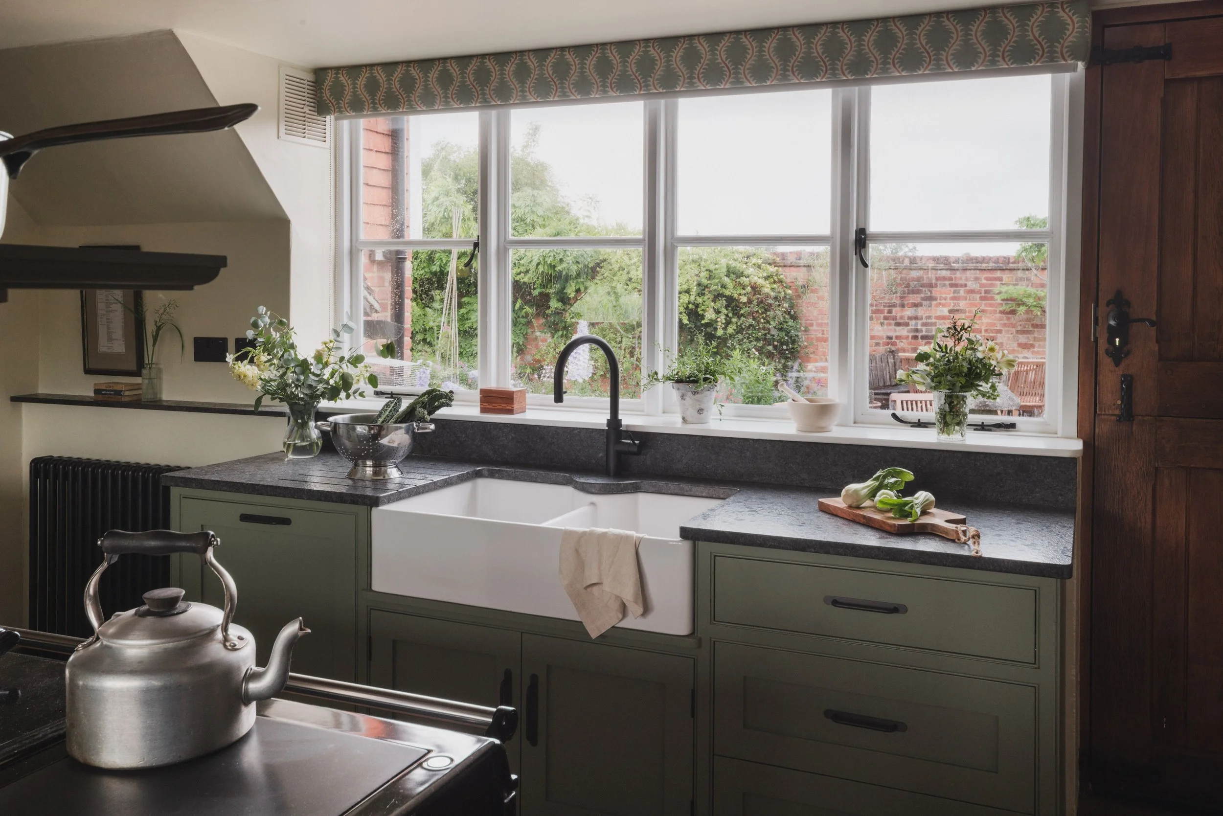 A kitchen with a large window above the sink, decorated with potted plants, a wooden spice box, and a cutting board with vegetables. There is a tea kettle on the counter in the foreground.
