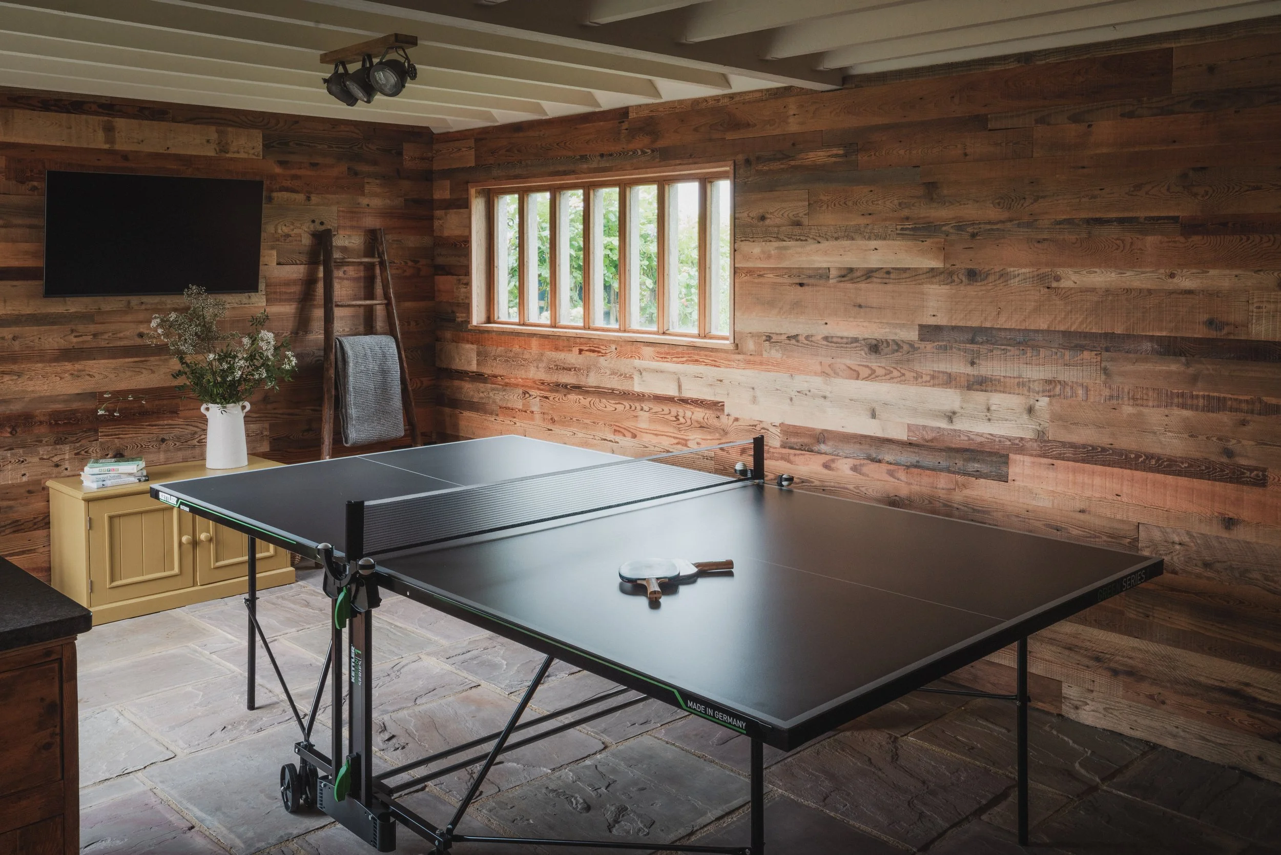 A rustic room with wooden panel walls, a window showing greenery outside, a flat-screen TV on the wall, a ladder, a table with a vase of flowers, a stack of books, and a ping pong table with paddles and balls.