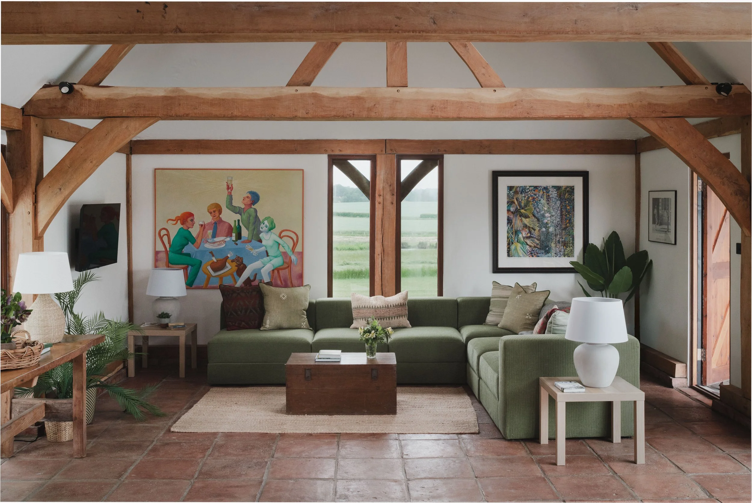 Living room with a green sectional sofa, artwork on the wall, and large windows showing a green landscape outside, with wooden beams and a terracotta tiled floor.