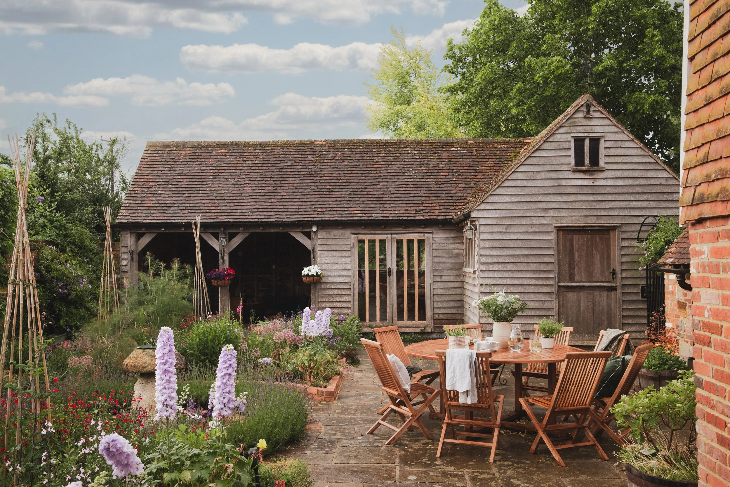 A rustic outdoor garden patio with a wooden barn-style shed, surrounded by colorful flowers and lush green trees, with a patio table and chairs set for dining.