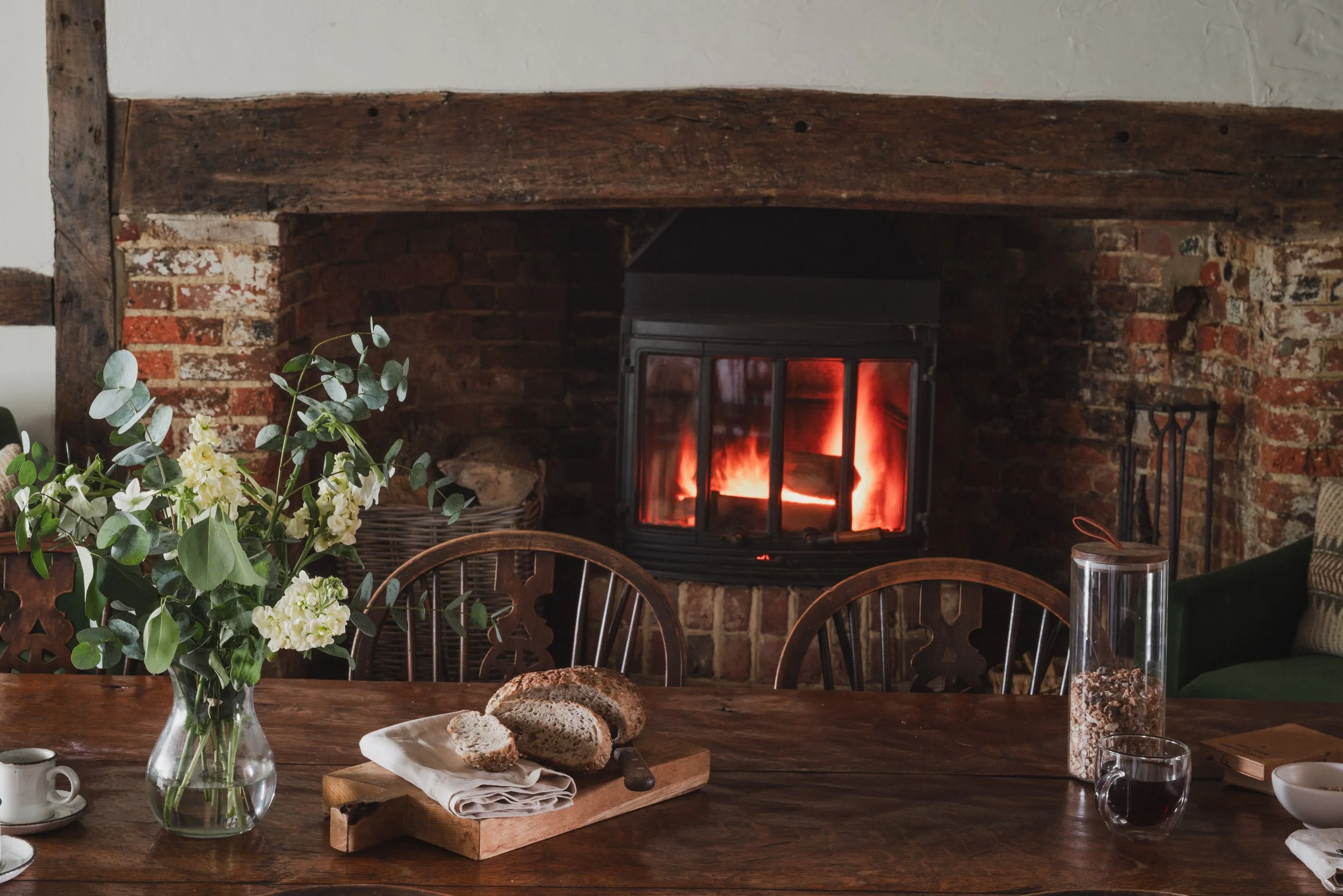 A cozy room with a wood-burning stove fireplace, a wooden dining table with a vase of white flowers, slices of bread on a cutting board, and chairs, with a brick and wood mantel surrounding the fireplace.