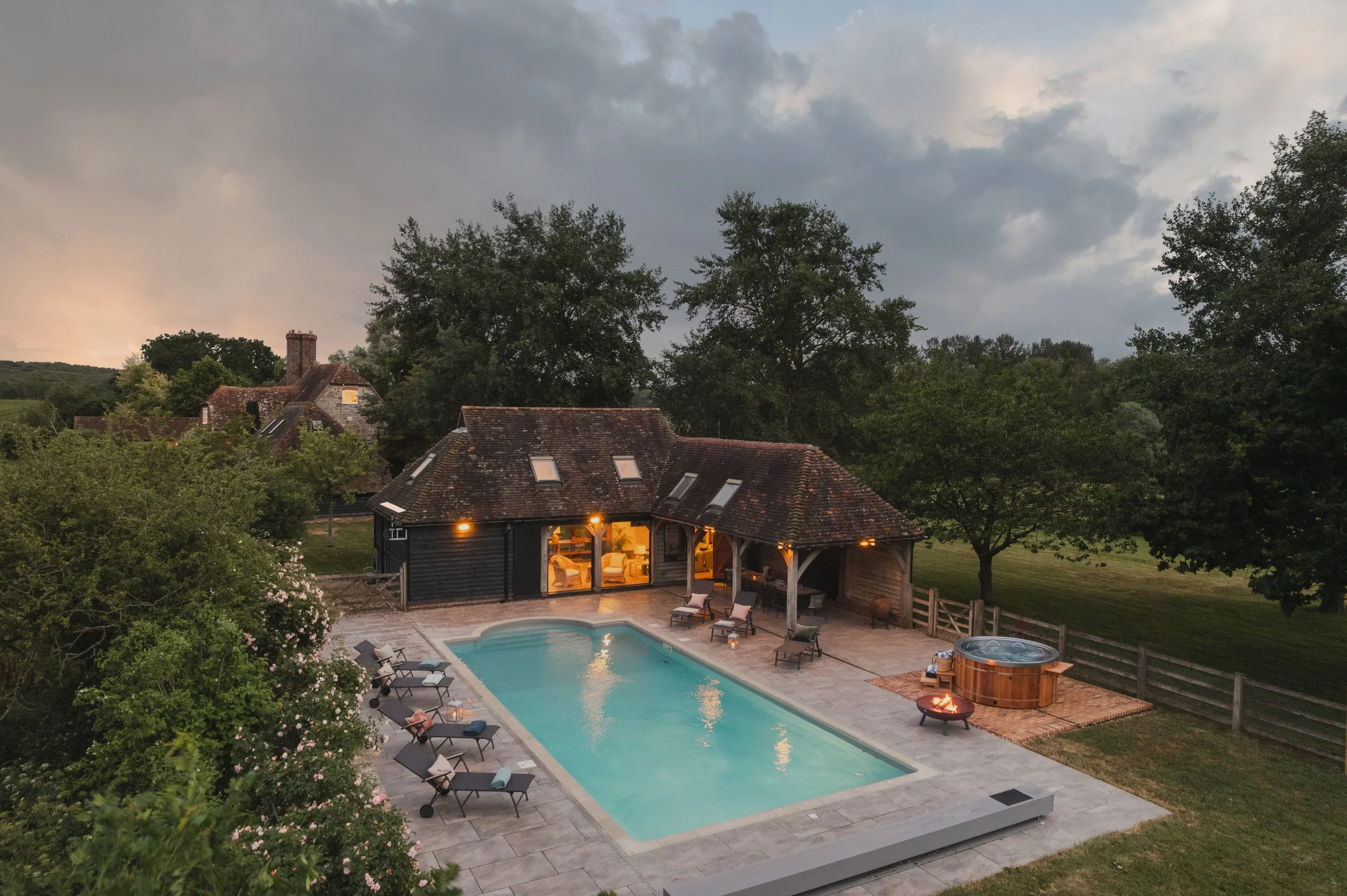 A backyard scene at dusk with a swimming pool, lounge chairs, and a hot tub near a house with large windows. The house has a covered porch with seating, and the background features trees, a cloudy sky, and nearby houses with chimneys. The outdoor are