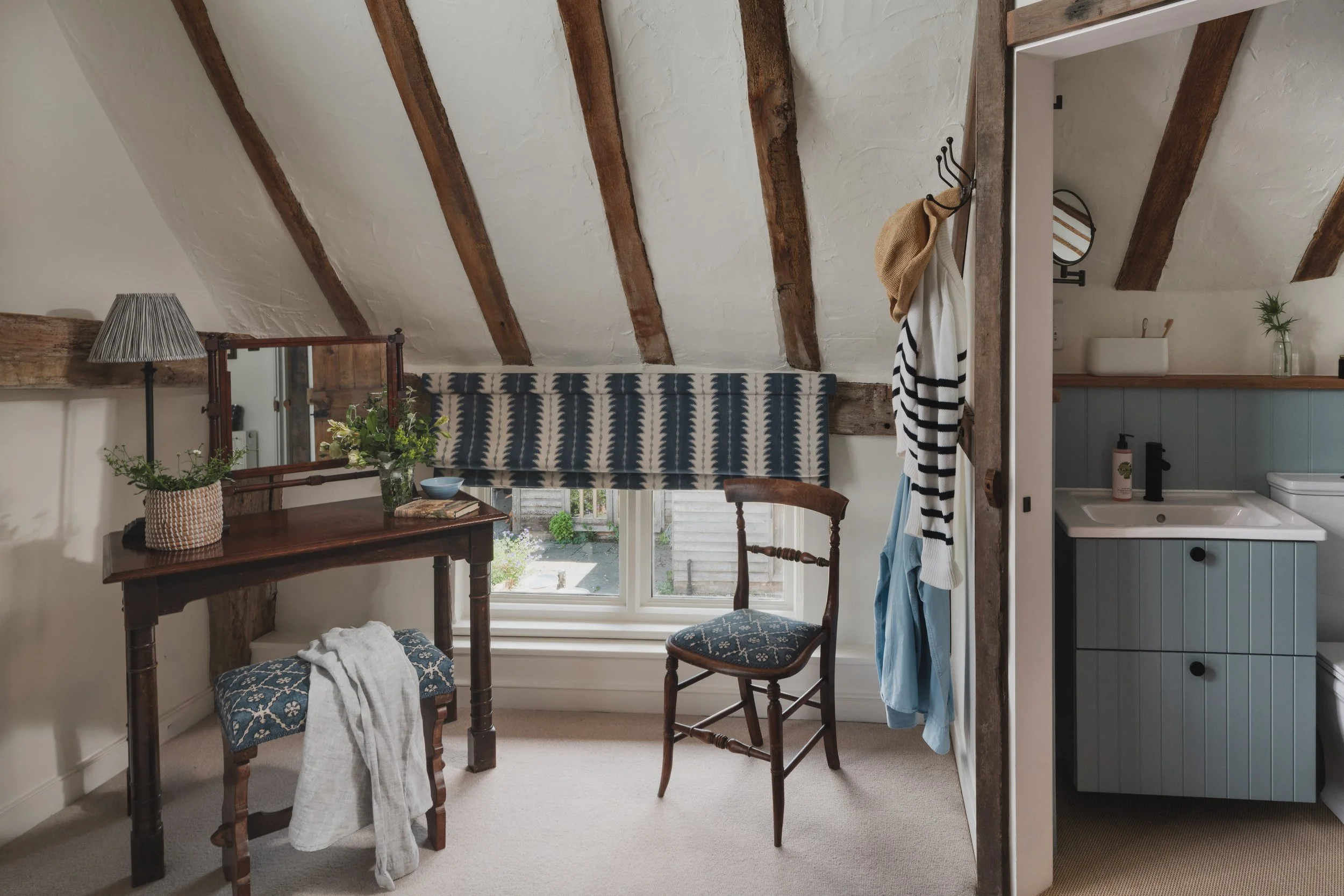 The image depicts a cozy room with a sloped ceiling supported by exposed wooden beams. There is a window with a view of a garden, a small wooden table with a vase of flowers, and three chairs with patterned upholstery. To the right, a partial view of