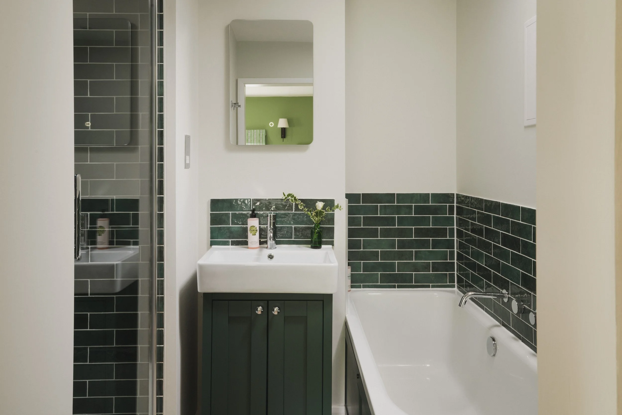 Bathroom with green tiled lower walls, white upper walls, a bathtub, a vanity with a green cabinet, a mirror, a small plant, and toiletries.